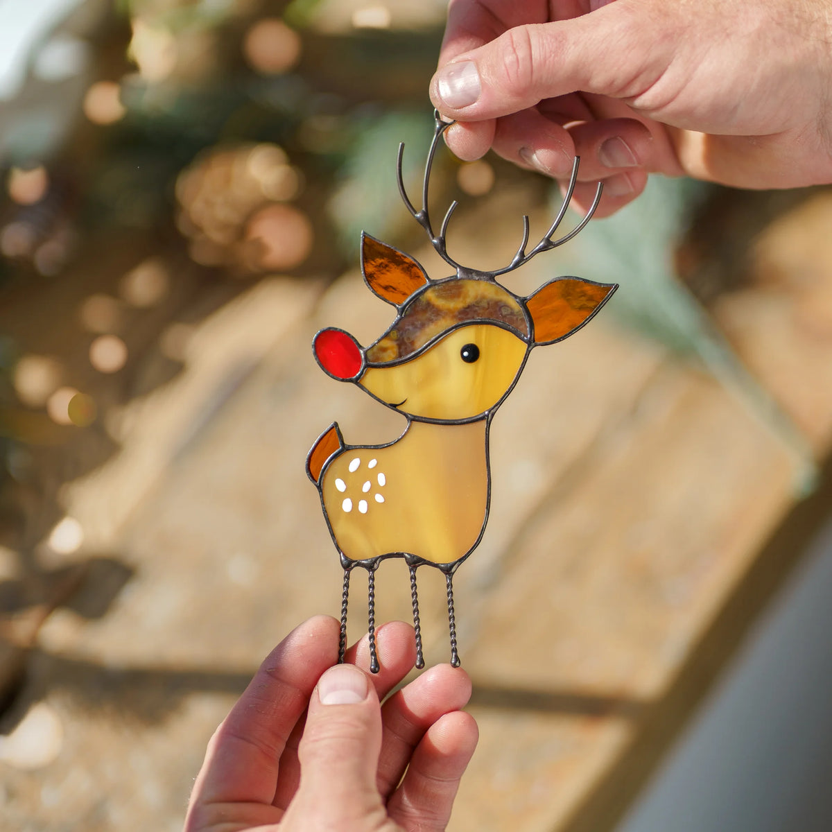 Two hands holding a stained glass reindeer suncatcher outdoors near pinecones and soft pine branches in warm golden sunlight.
