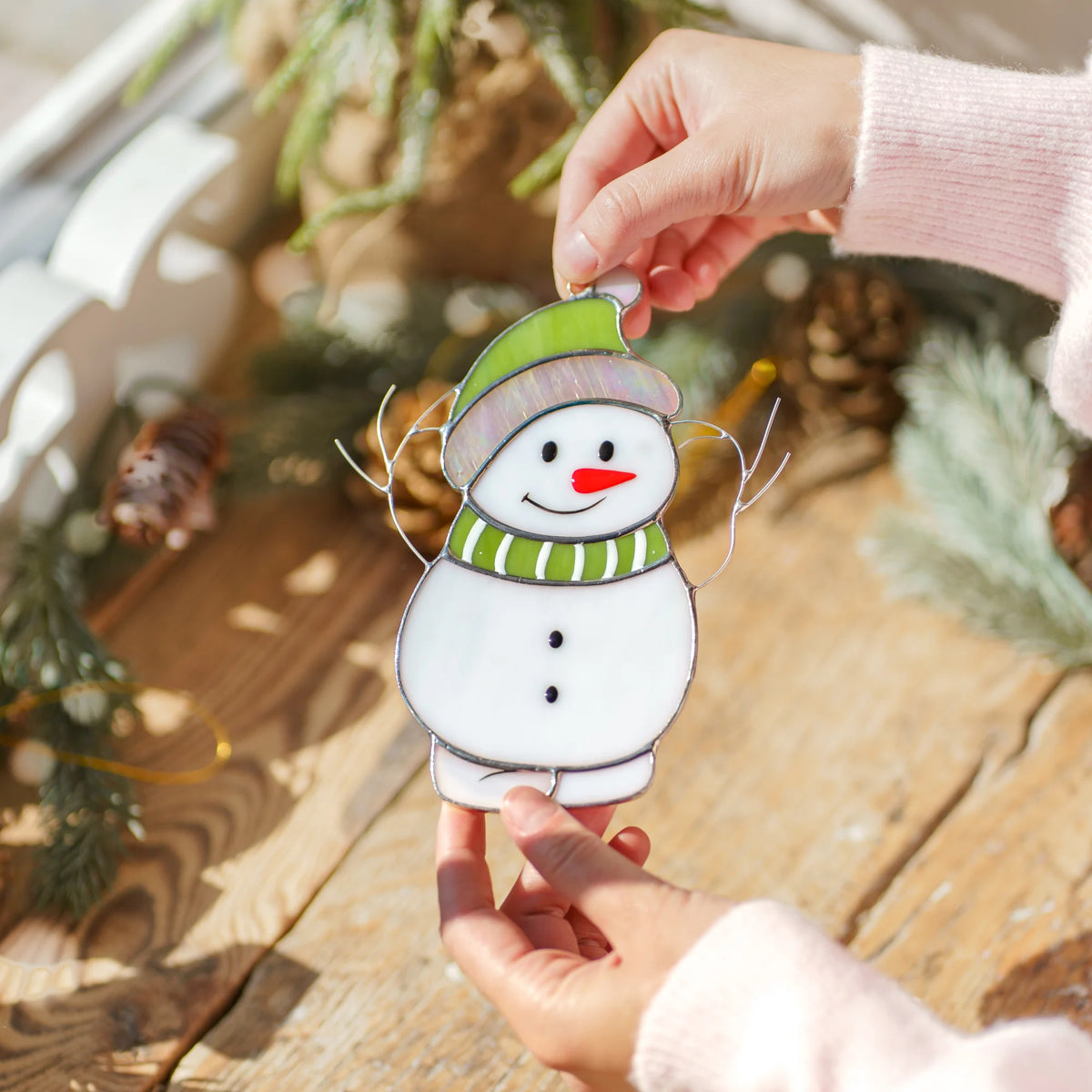 Stained glass snowman suncatcher held above a rustic wooden table decorated with pinecones and fresh pine branches outdoors.