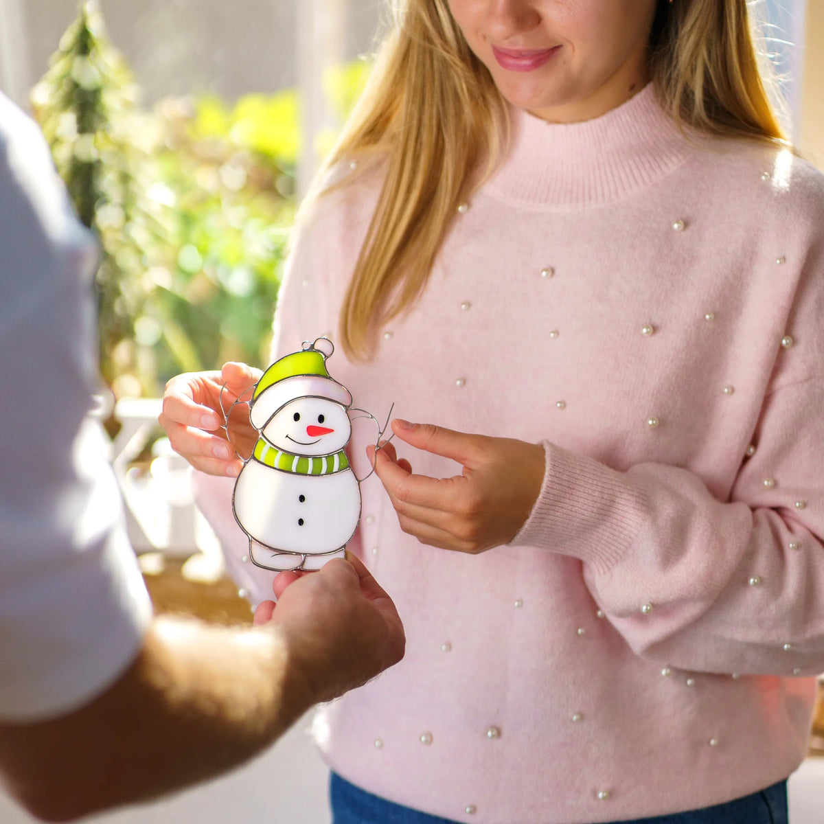 Two people exchanging a stained glass snowman suncatcher by hand, one wearing a pink pearl-studded sweater, in soft sunlight.