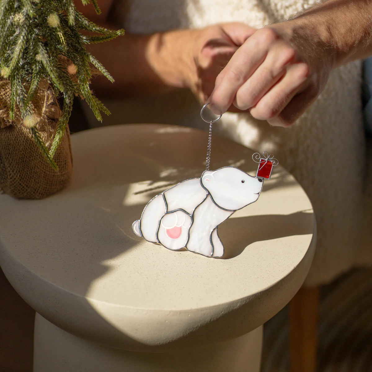 Sitting polar bear with a Christmas gift stained glass suncatcher held above a round side table in the sun, a pink heart on its paw - a sweet handmade Christmas ornament