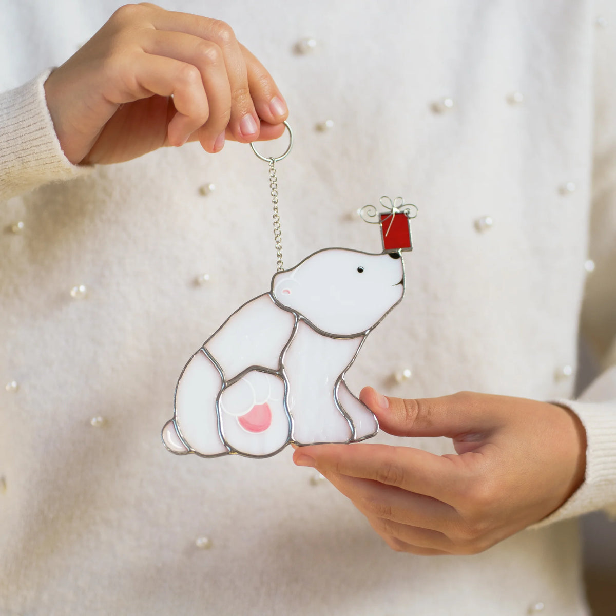 Hands holding the polar bear with a Christmas gift stained glass suncatcher against a white sweater, a tiny red gift box on its nose - handmade Christmas ornament and holiday gift