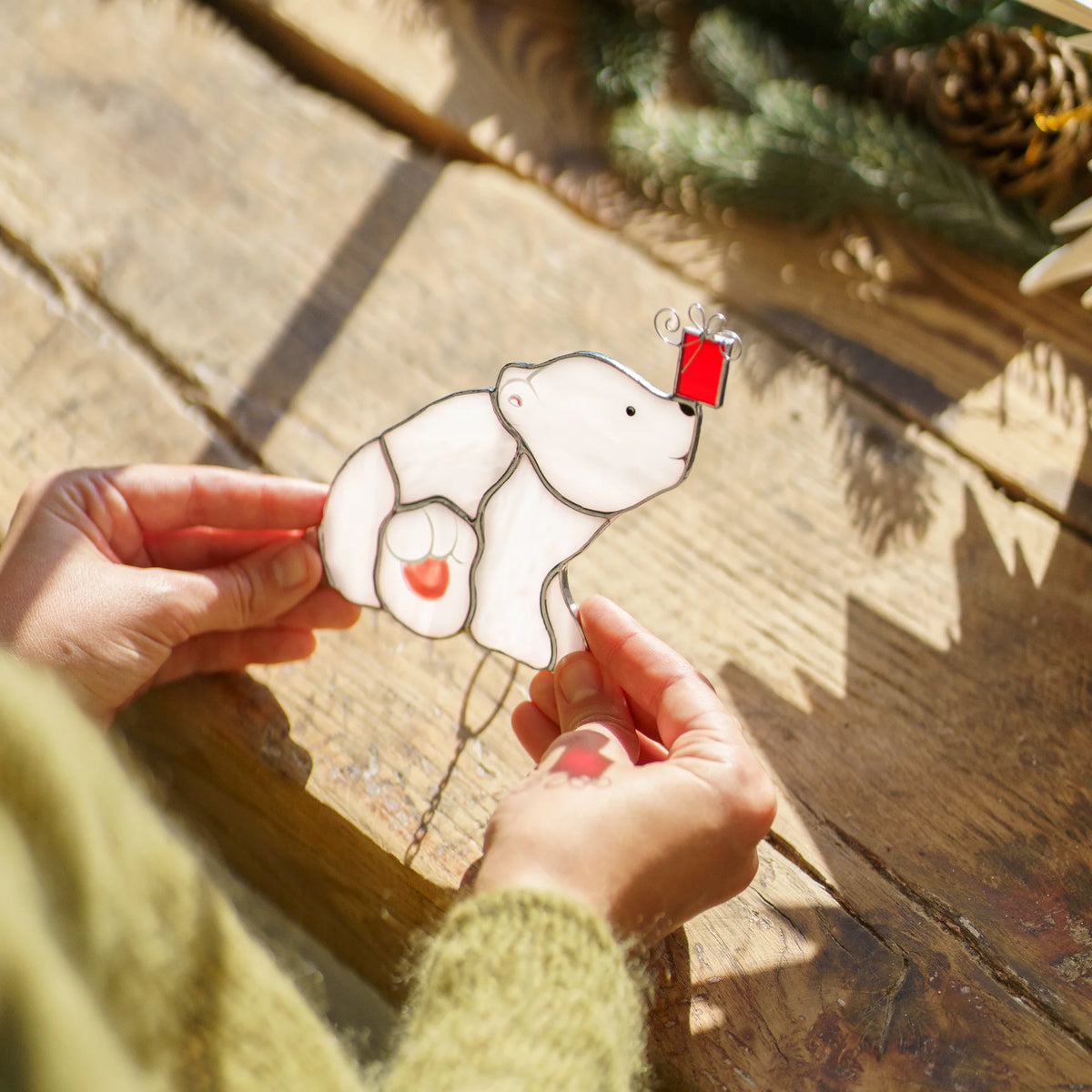 Close-up of the polar bear with a Christmas gift stained glass ornament over a rustic wooden sill with pine cones and fir in the sun - handmade Christmas stained glass suncatcher