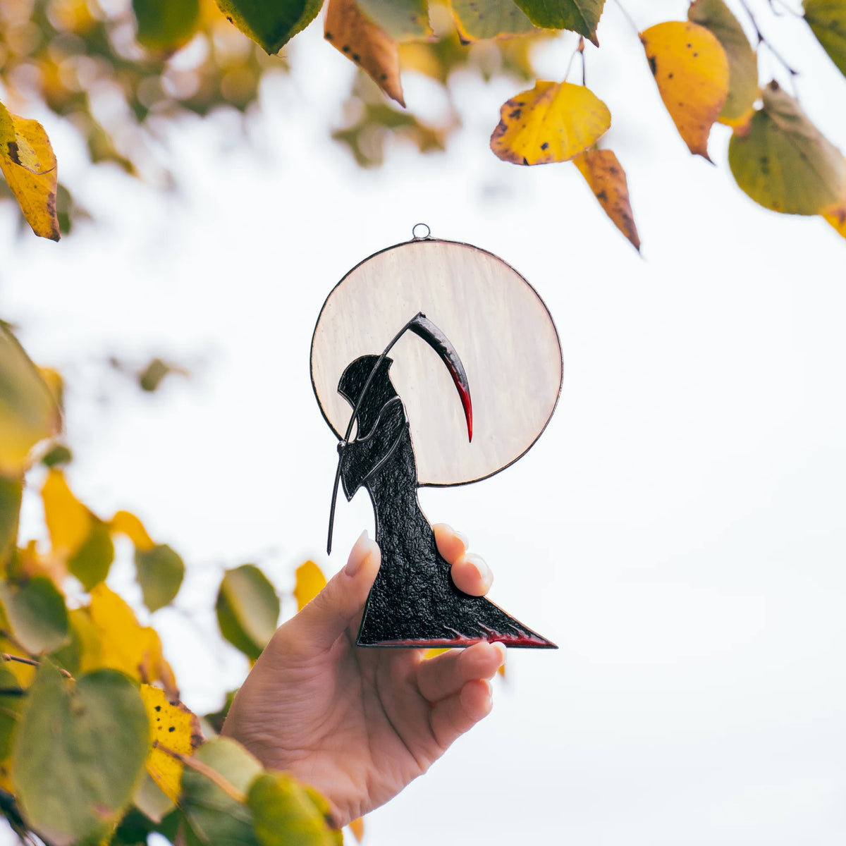 Hand holding the Grim Reaper Halloween stained glass window hanging under a branch of yellow autumn leaves - handmade gothic decor for a window through the season