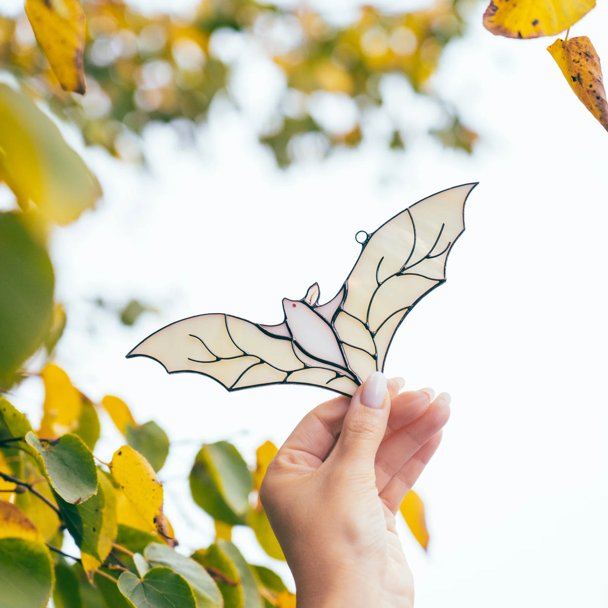 Hand raising the white stained glass bat suncatcher toward golden autumn leaves and branches against a bright overcast sky.