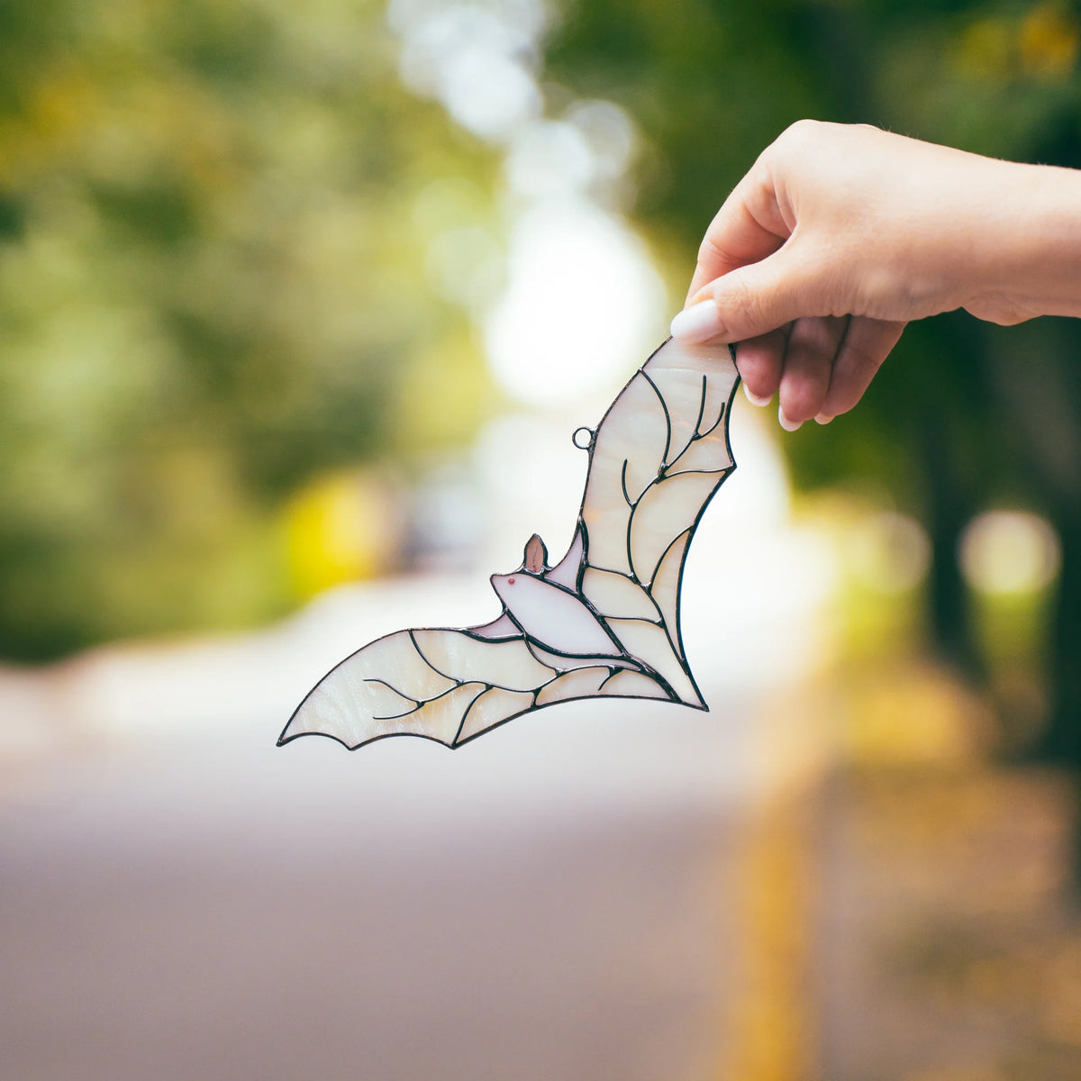Two open hands cupping the white stained glass bat suncatcher, framed by warm orange and yellow autumn foliage in the background.
