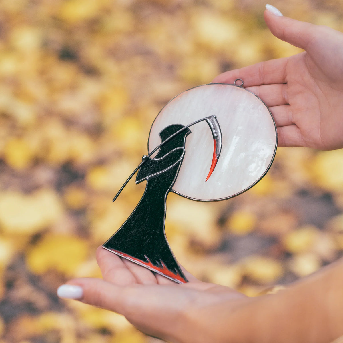 Grim Reaper stained glass window hanging held in a hand above fallen yellow leaves, showing the textured black robe, red scythe edge and pale opal moon