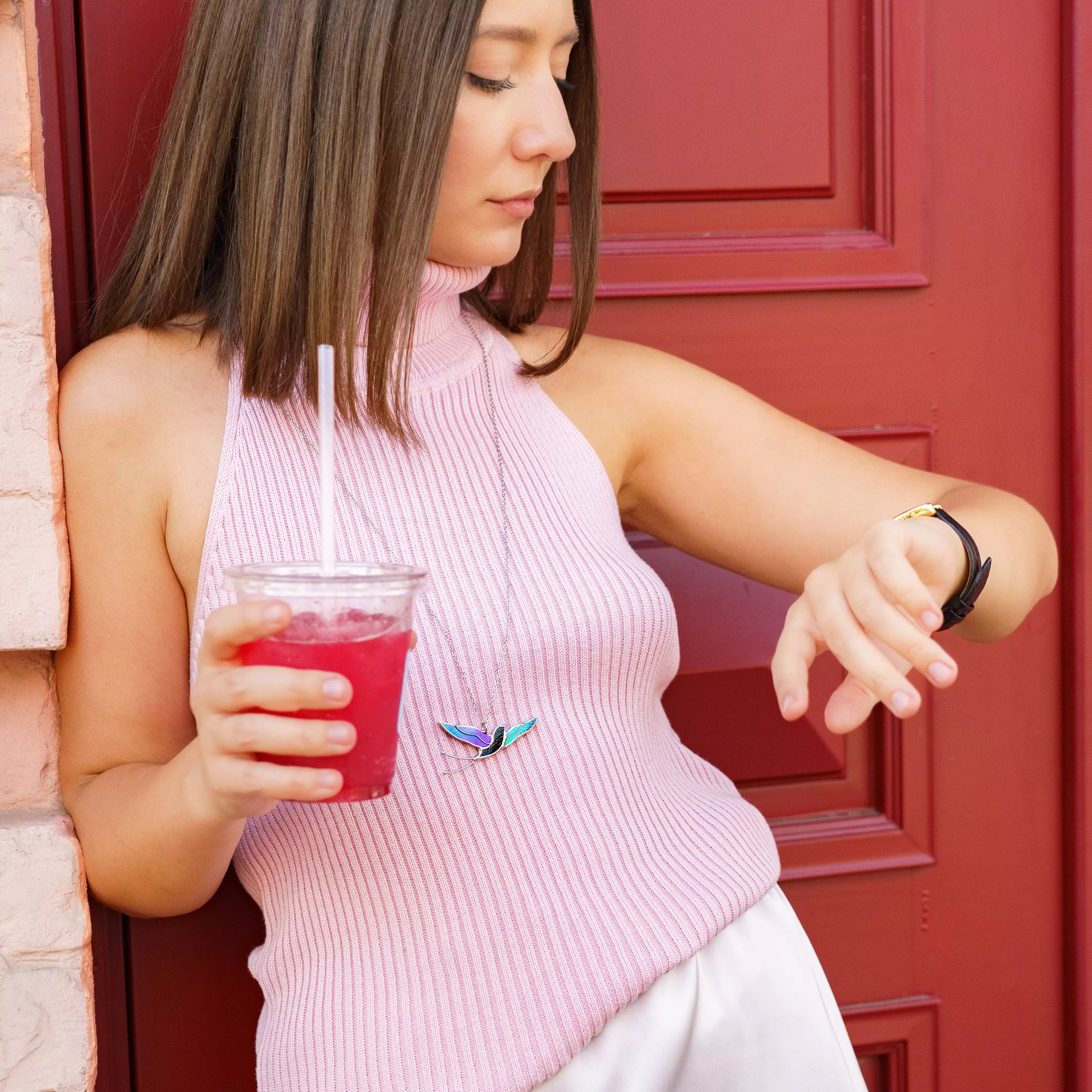 Woman in a pink halter top by a red door wearing the stained glass swallow necklace on a long silver chain, the iridescent bird pendant in flight at her chest - handmade bird lover jewelry