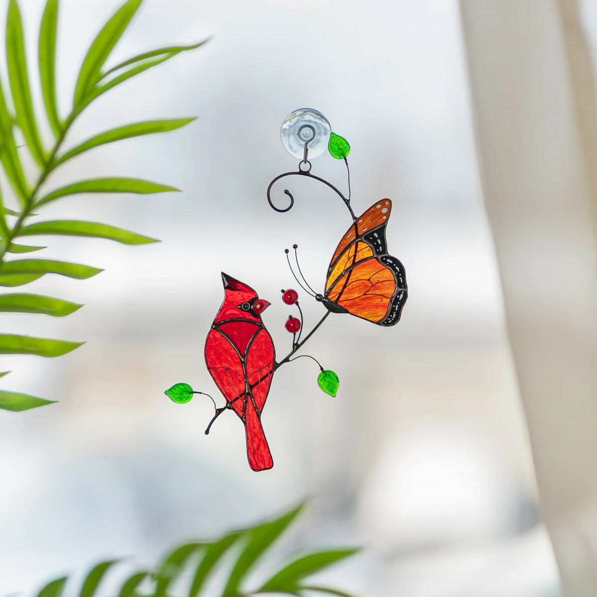 Cardinal and monarch butterfly stained glass suncatcher hanging by a suction cup on a bright window beside palm leaves - handmade red cardinal and orange butterfly window decor