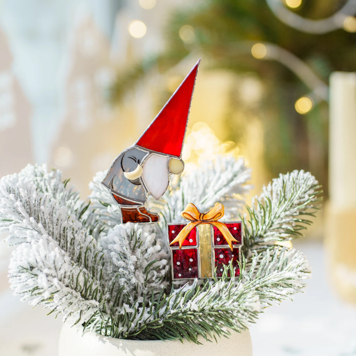 Close-up of the Christmas gnome with gift stained glass plant stakes among frosted fir branches with soft golden bokeh - handmade stained glass Christmas plant decor and holiday gift