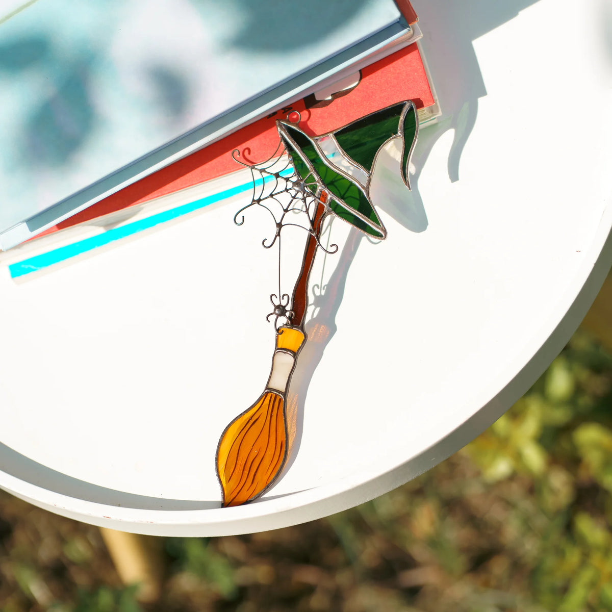 Handmade witch broom stained glass suncatcher lying on a white table beside a stack of books in bright sunlight, the green witch hat and amber bristles glowing through