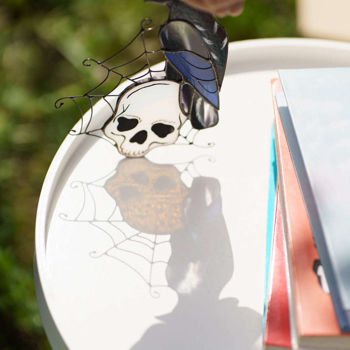 Close-up of the white glass skull and wire spider web of a handmade raven suncatcher in sunlight casting a soft shadow - gothic stained glass detail with painted bone shading for Halloween