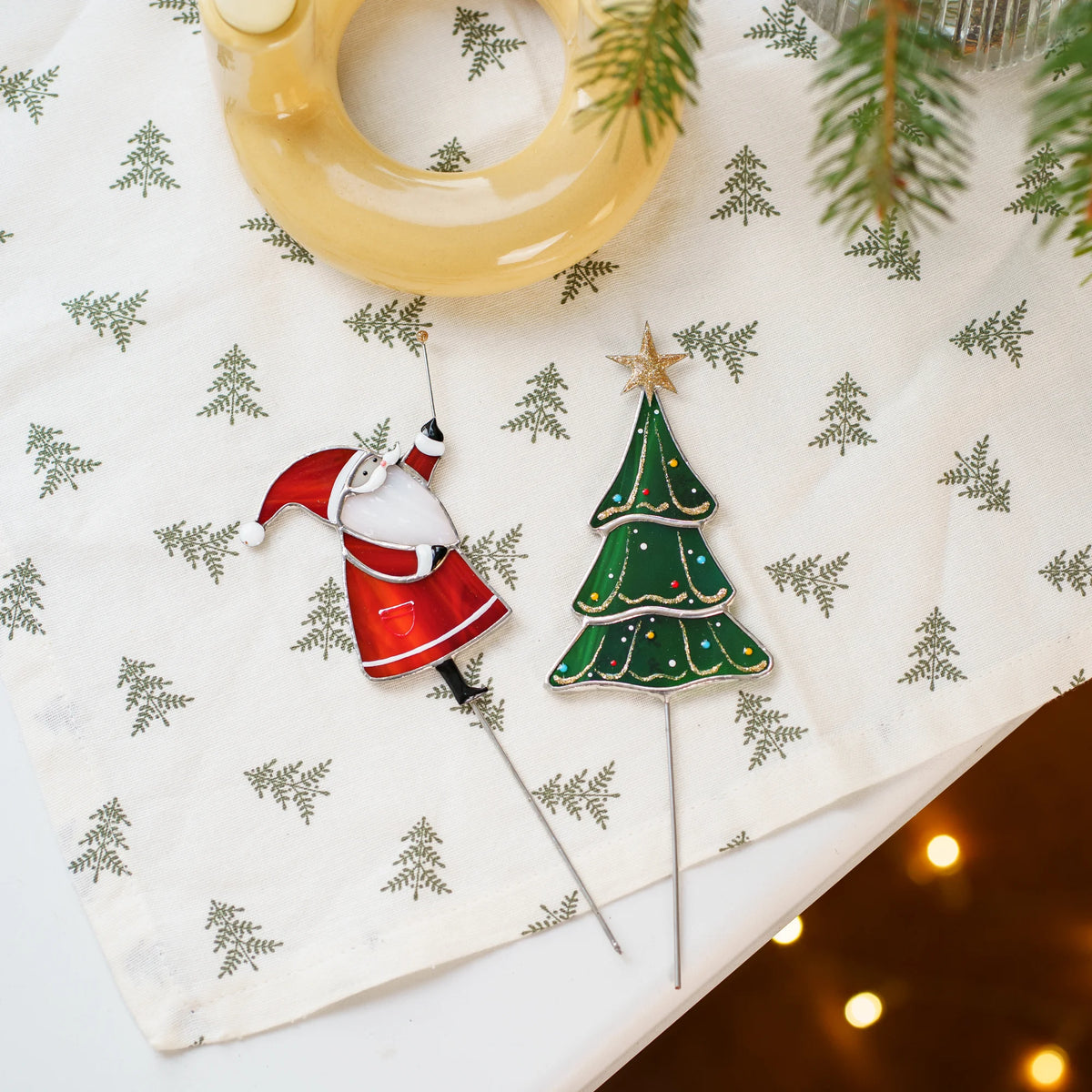 Santa and Christmas tree stained glass plant stakes lying on a white tablecloth with tiny tree prints next to a wreath and fairy lights - handmade Christmas stained glass ornaments
