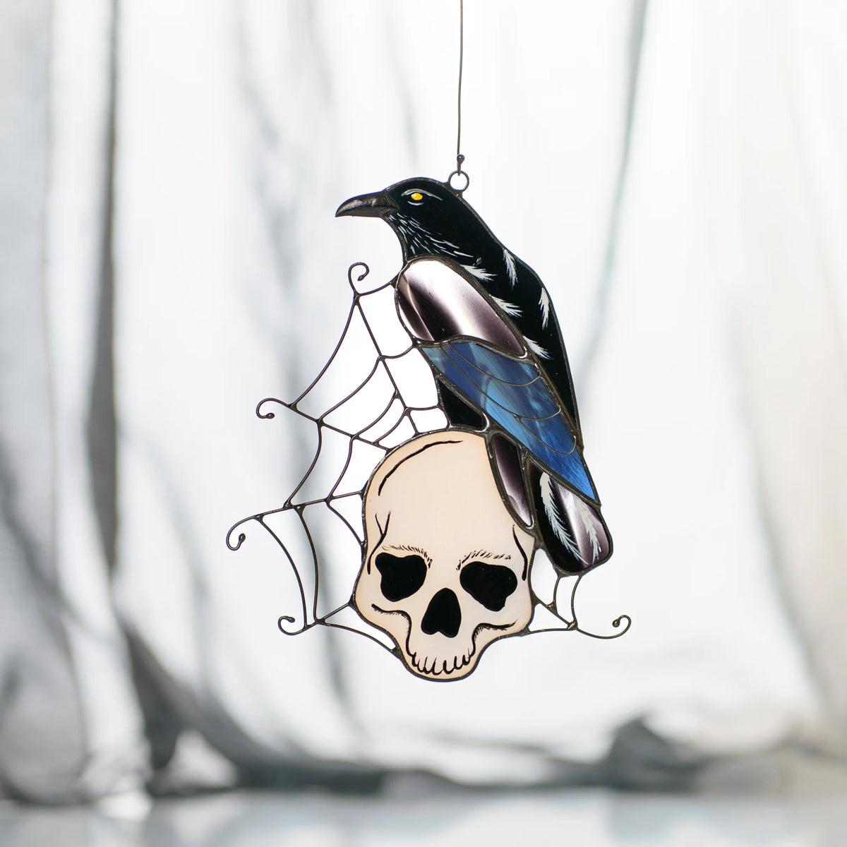 Stained glass raven suncatcher on a bone-white skull in a wire spider web against soft grey drapery, iridescent blue and lilac wing over black plumage - handmade gothic Halloween window art