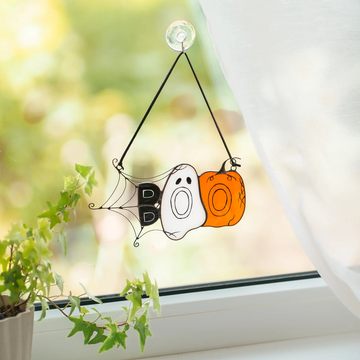 Handmade BOO Halloween stained glass suncatcher resting on a white table beside a stack of books, bright sun throwing an orange pumpkin shadow across the surface