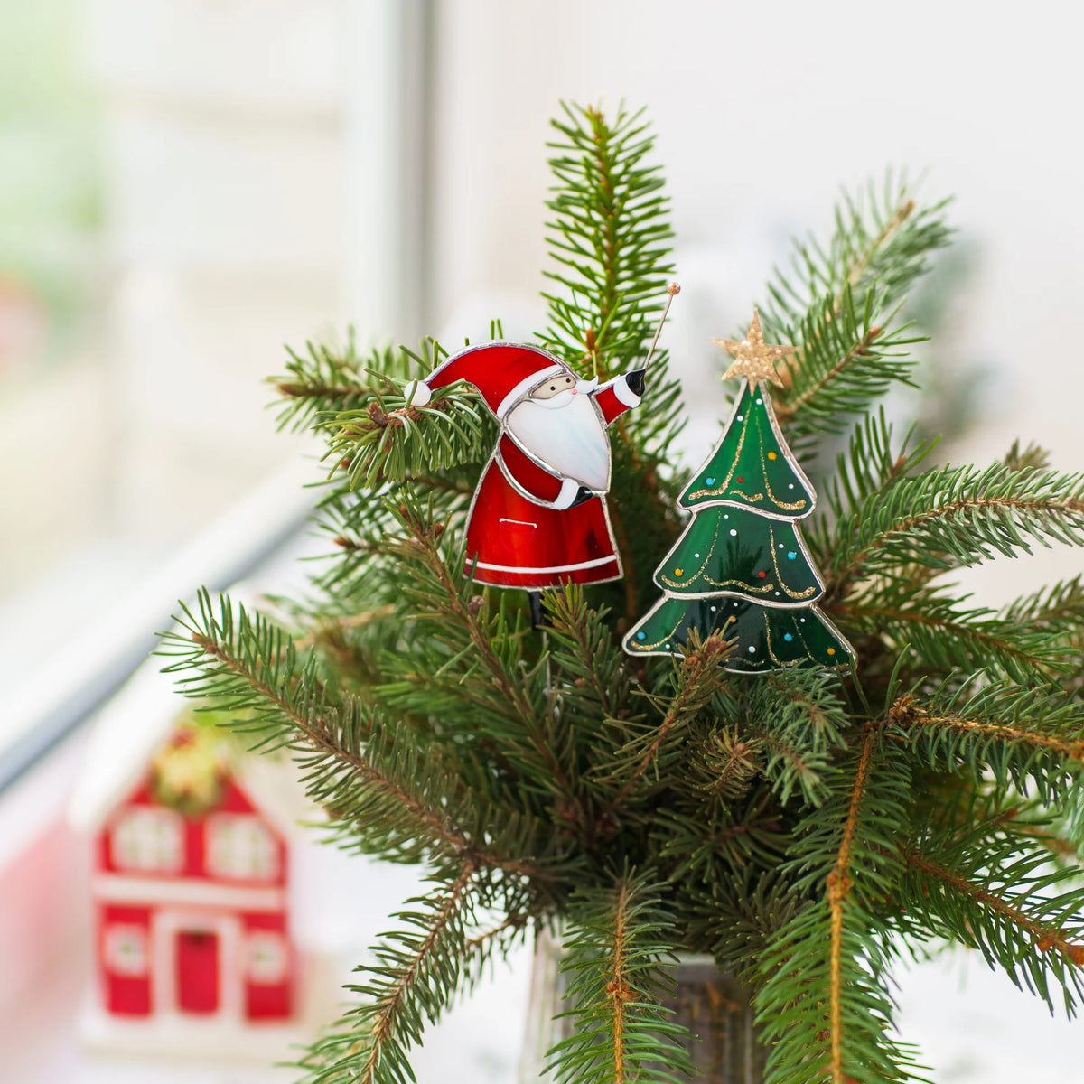 Santa and Christmas tree stained glass plant stakes tucked into fresh pine branches in a glass vase by a bright window - handmade Christmas stained glass decor for vases and planters