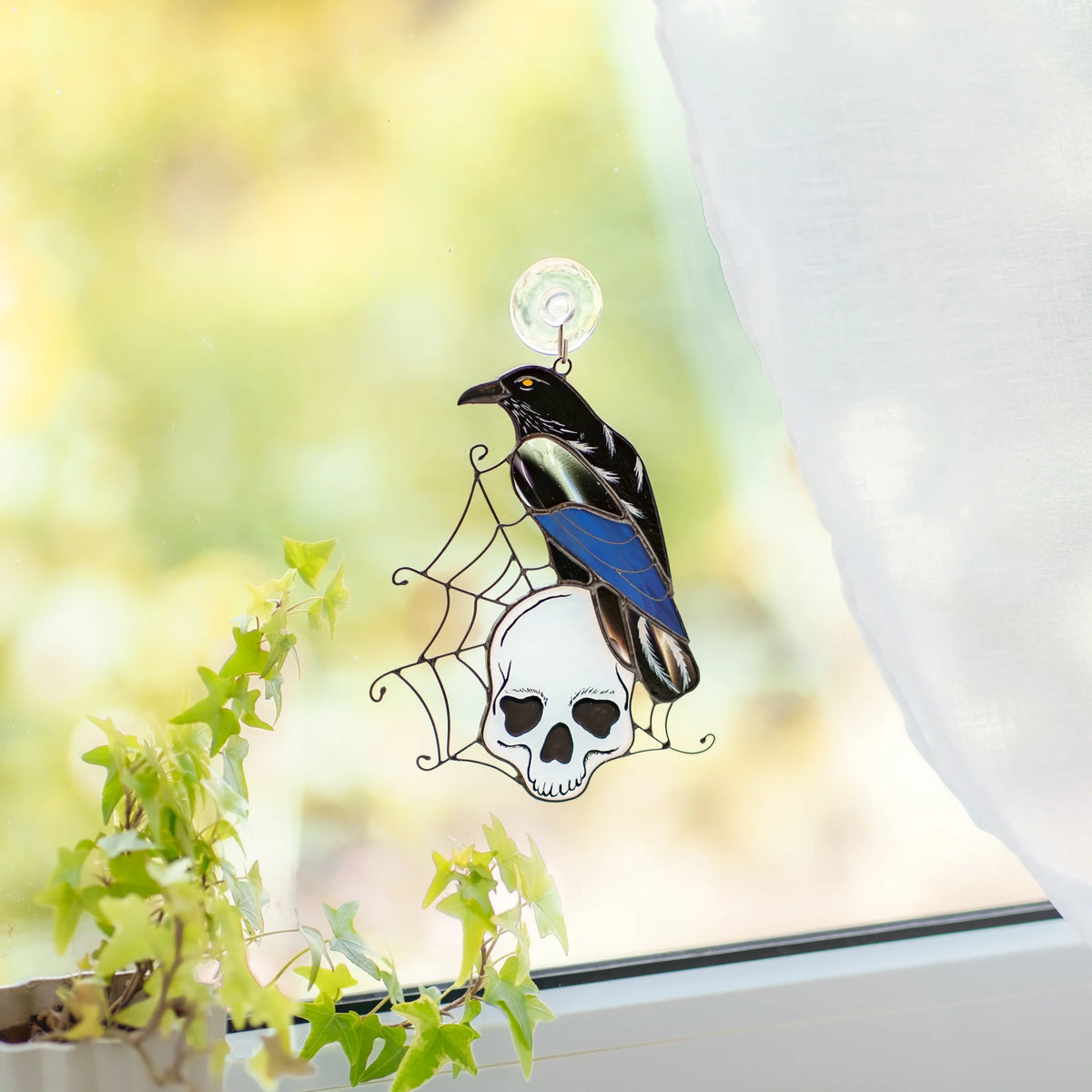 Raven on a skull stained glass suncatcher hanging on a window by a suction hook with ivy nearby, black raven and iridescent blue wing over a white skull - handmade gothic Halloween window decor