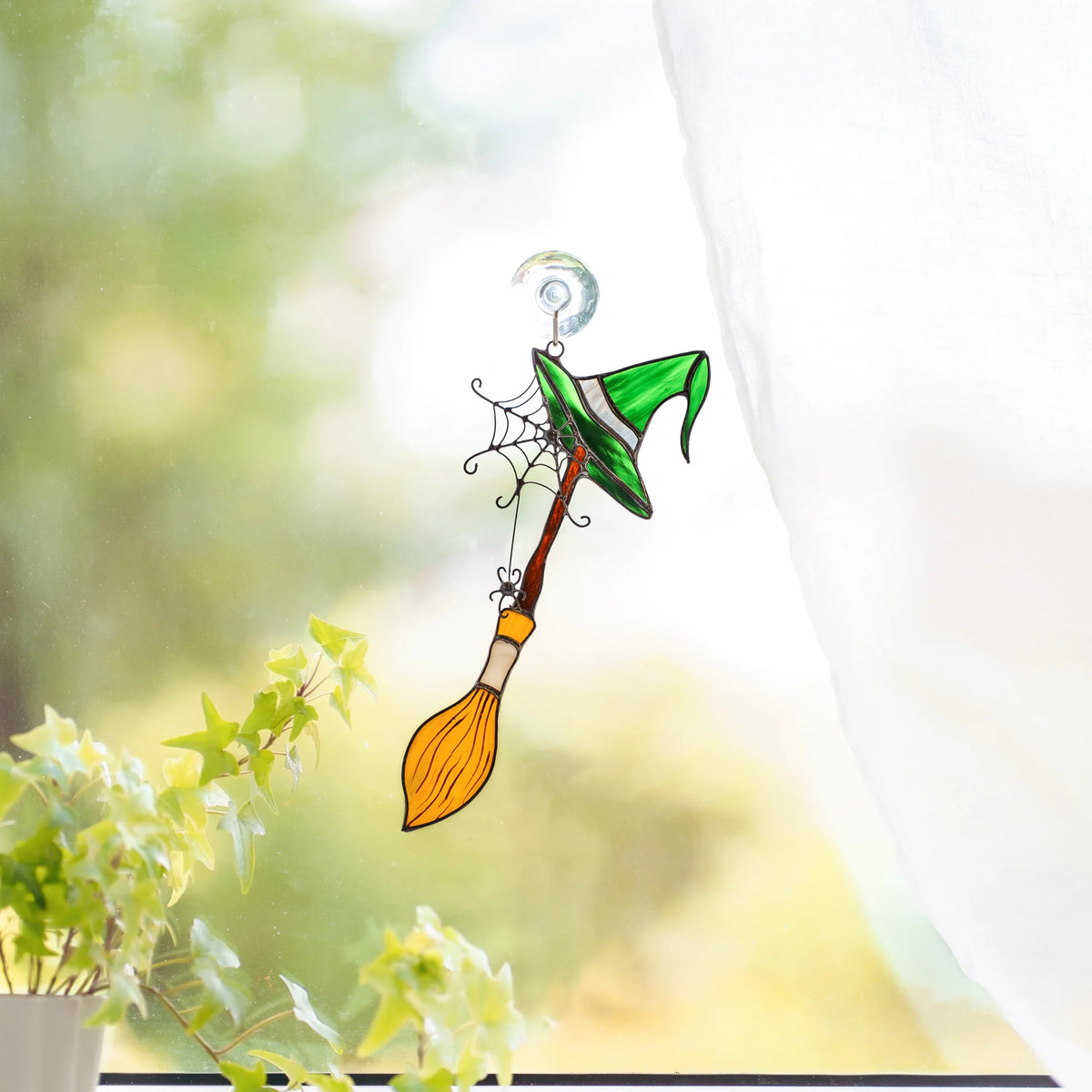 Witch broom and hat stained glass suncatcher hanging on a sunny window hook above an ivy plant - handmade Halloween window decor for a kitchen or living room