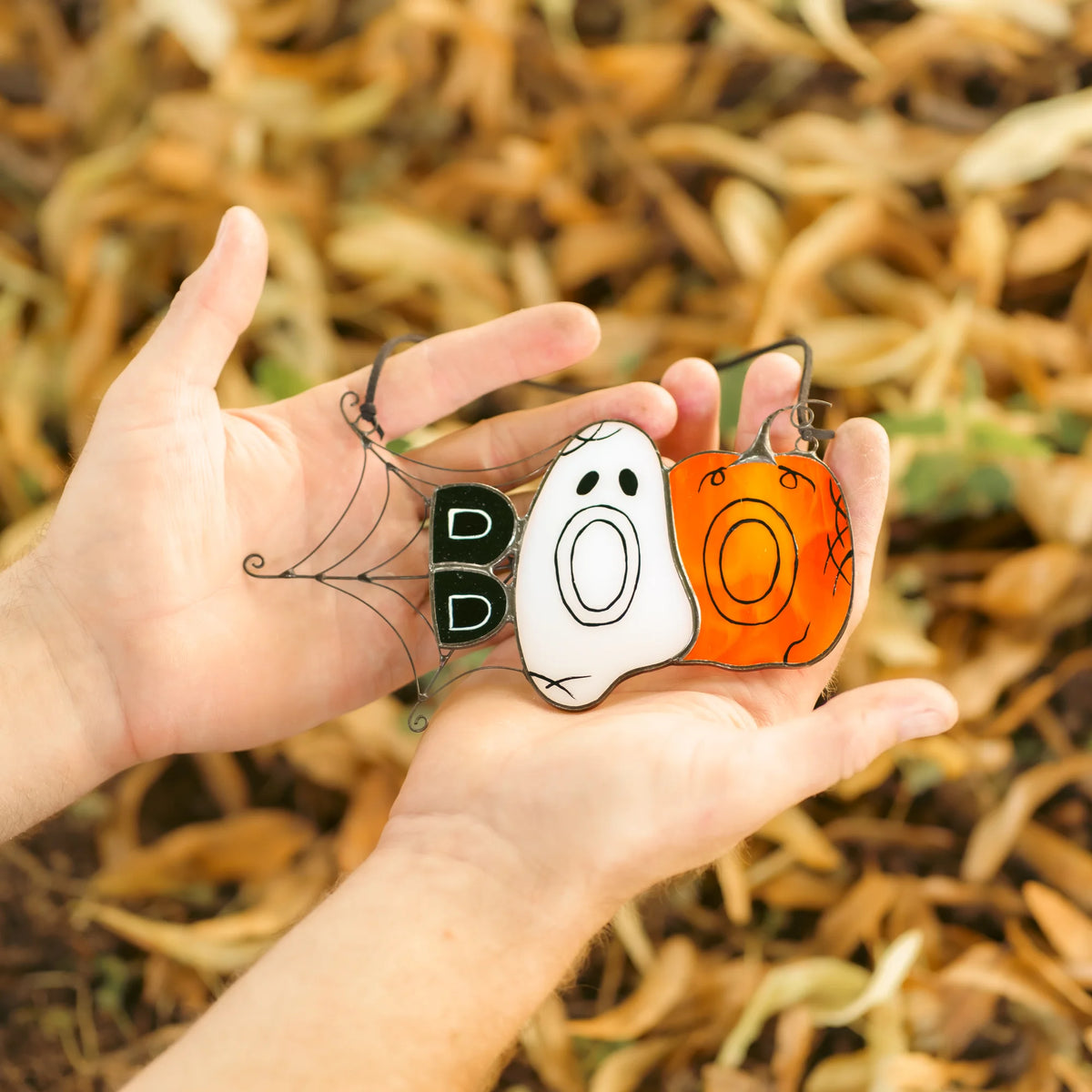 Hand holding the handmade BOO Halloween stained glass suncatcher by its cord against a green garden background, showing the ghost, pumpkin and wire spiderweb