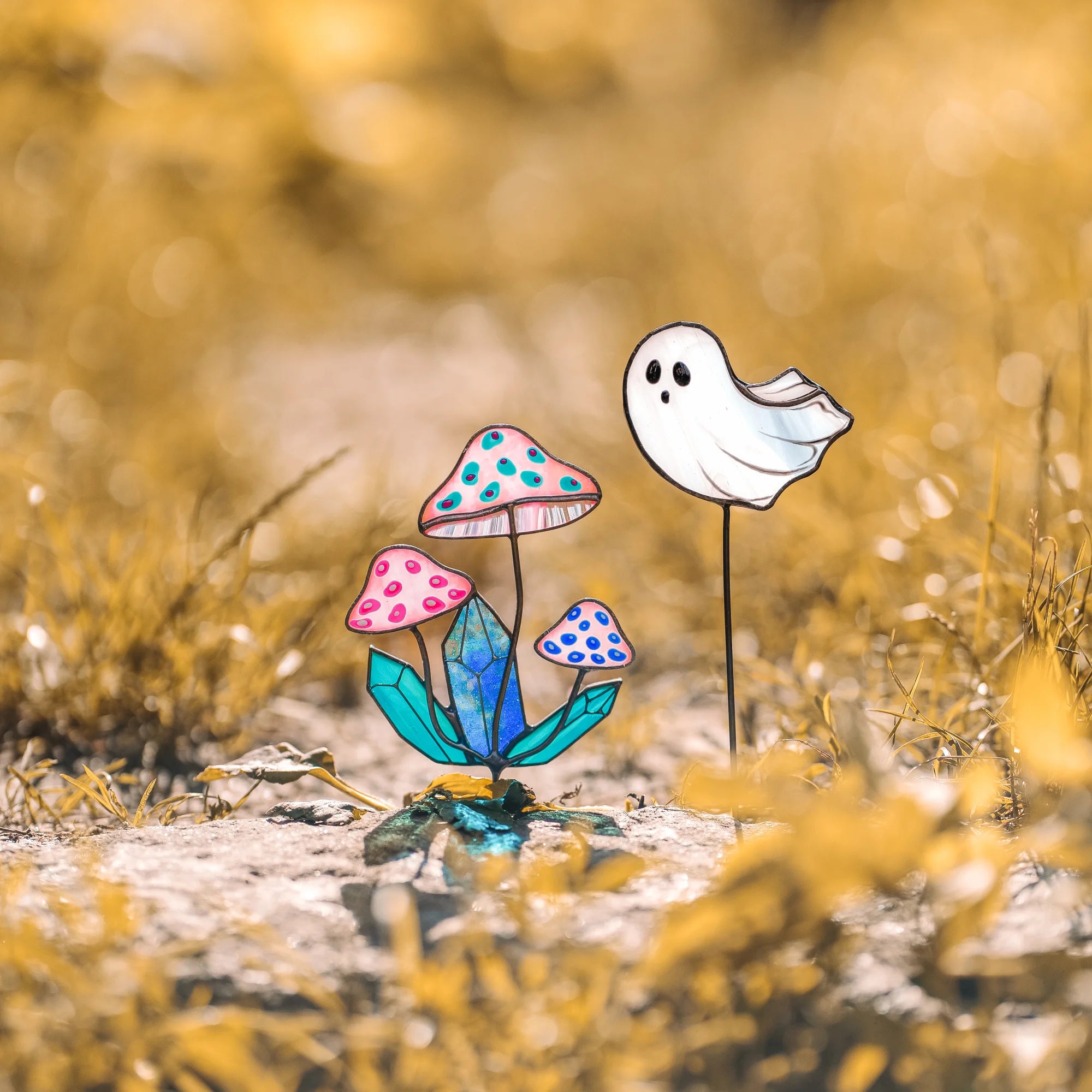 Mushroom and ghost stained glass garden art staked in golden autumn grass - pink iridescent toadstools around a blue crystal and a small white opalescent ghost catching the sun