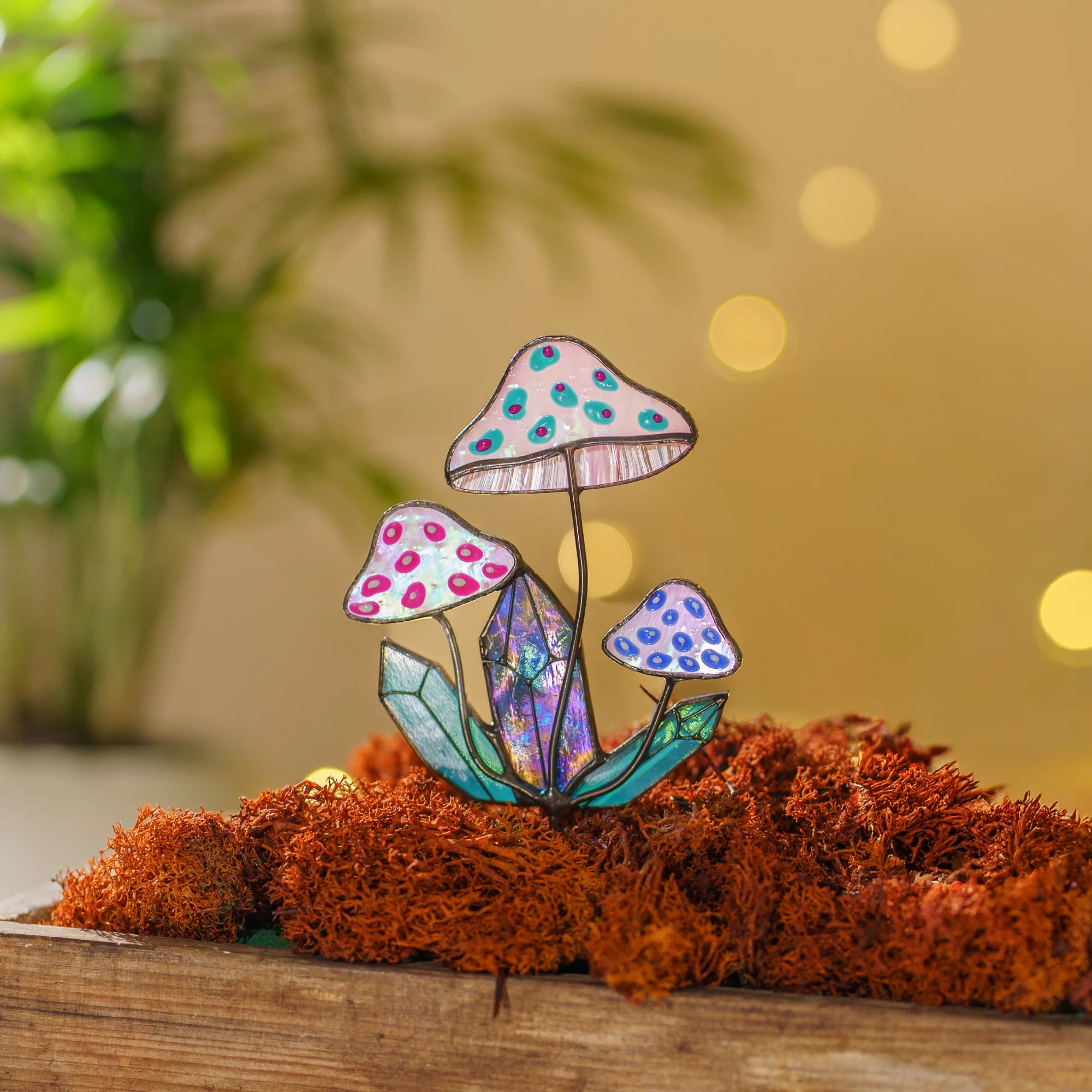 Close view of the iridescent stained glass mushroom plant stake in orange moss with warm bokeh lights - pink opal caps, teal leaves and a glowing blue-violet crystal