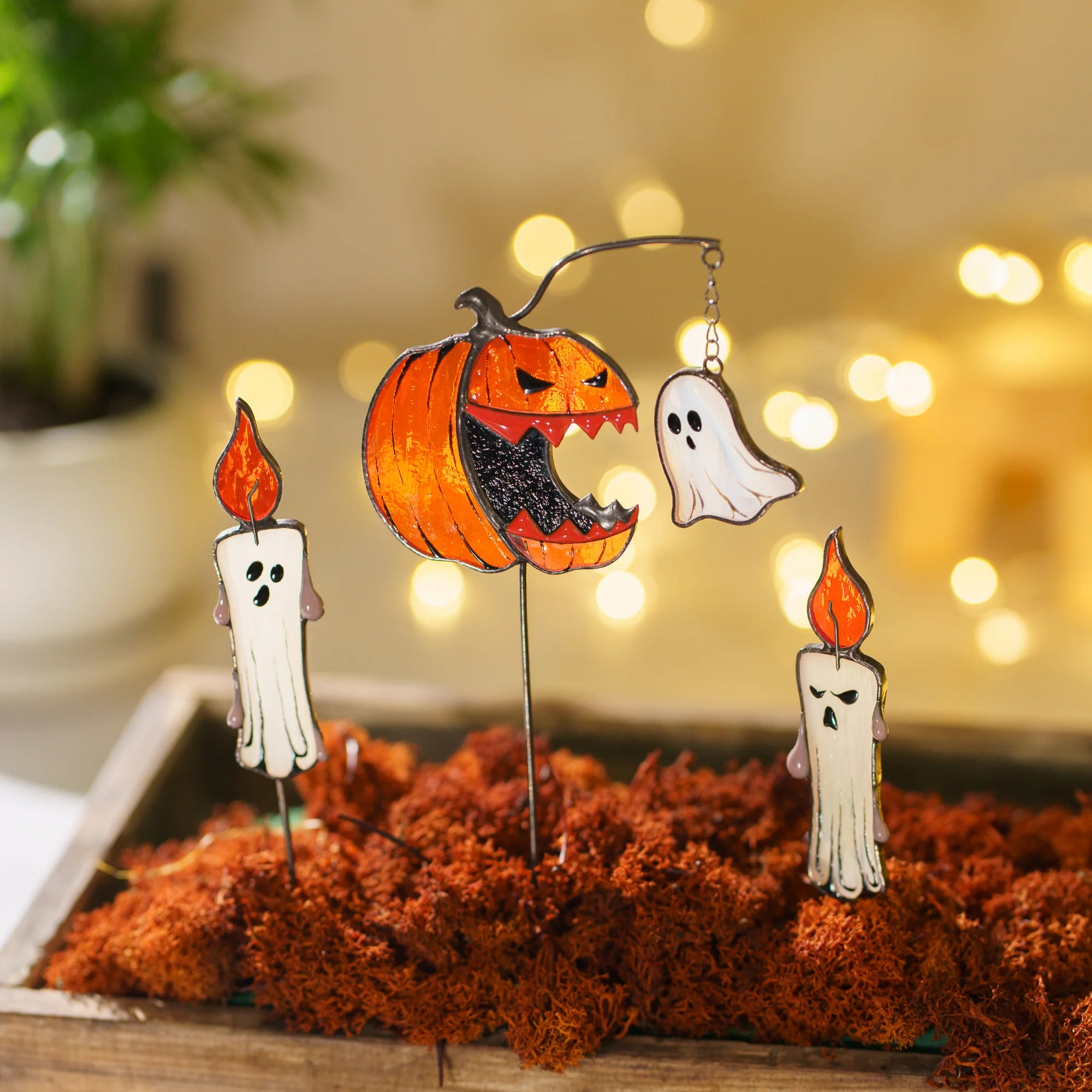 Halloween-themed stained glass plant stake decorations displayed in a tray of moss, featuring a carved jack-o'-lantern, two white candles with orange flames, and a white ghost, set against a warm blurred background with bokeh lights.