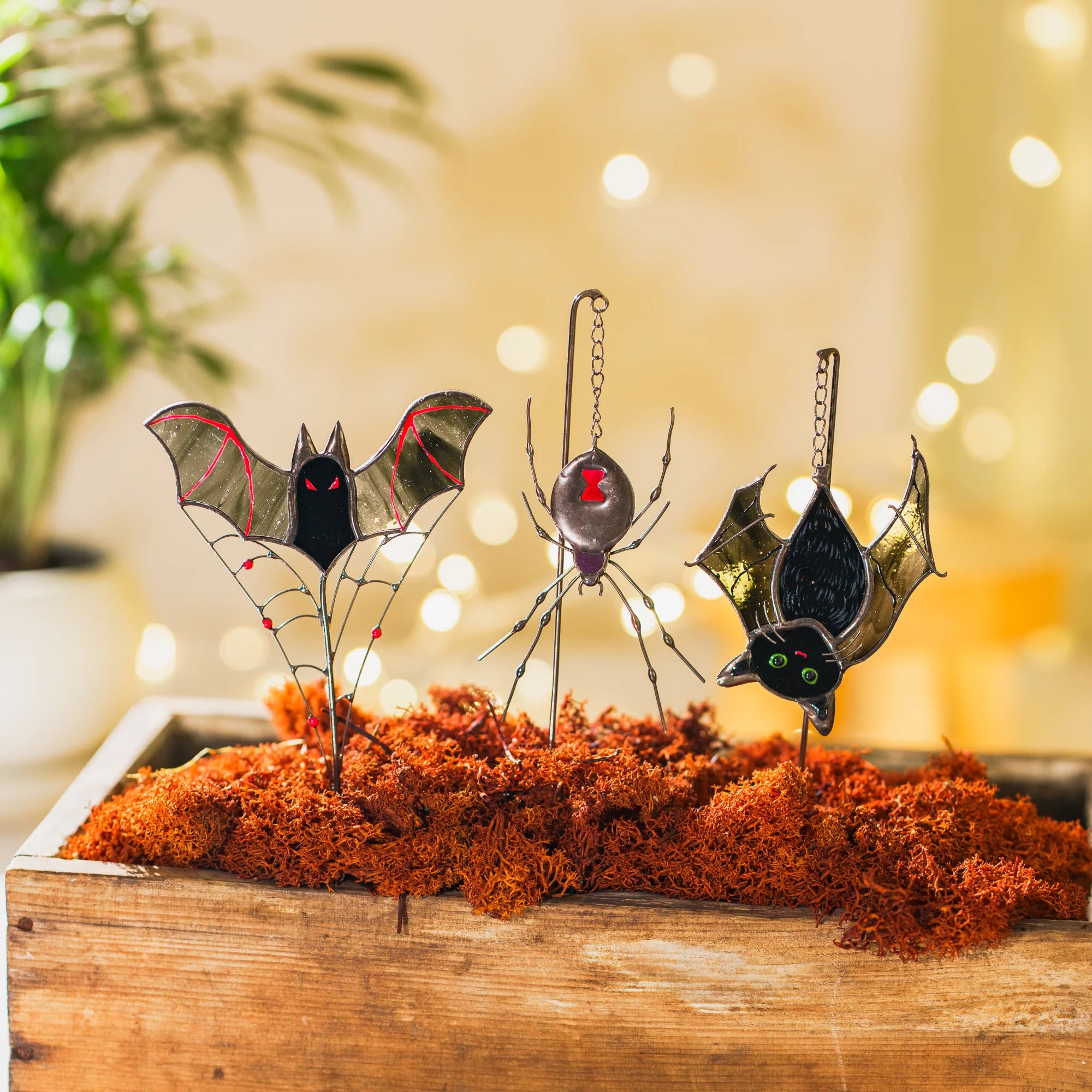 Bat and spider stained glass garden art staked in orange moss with warm bokeh lights behind - moody handmade Halloween decor for a cozy autumn corner or covered patio