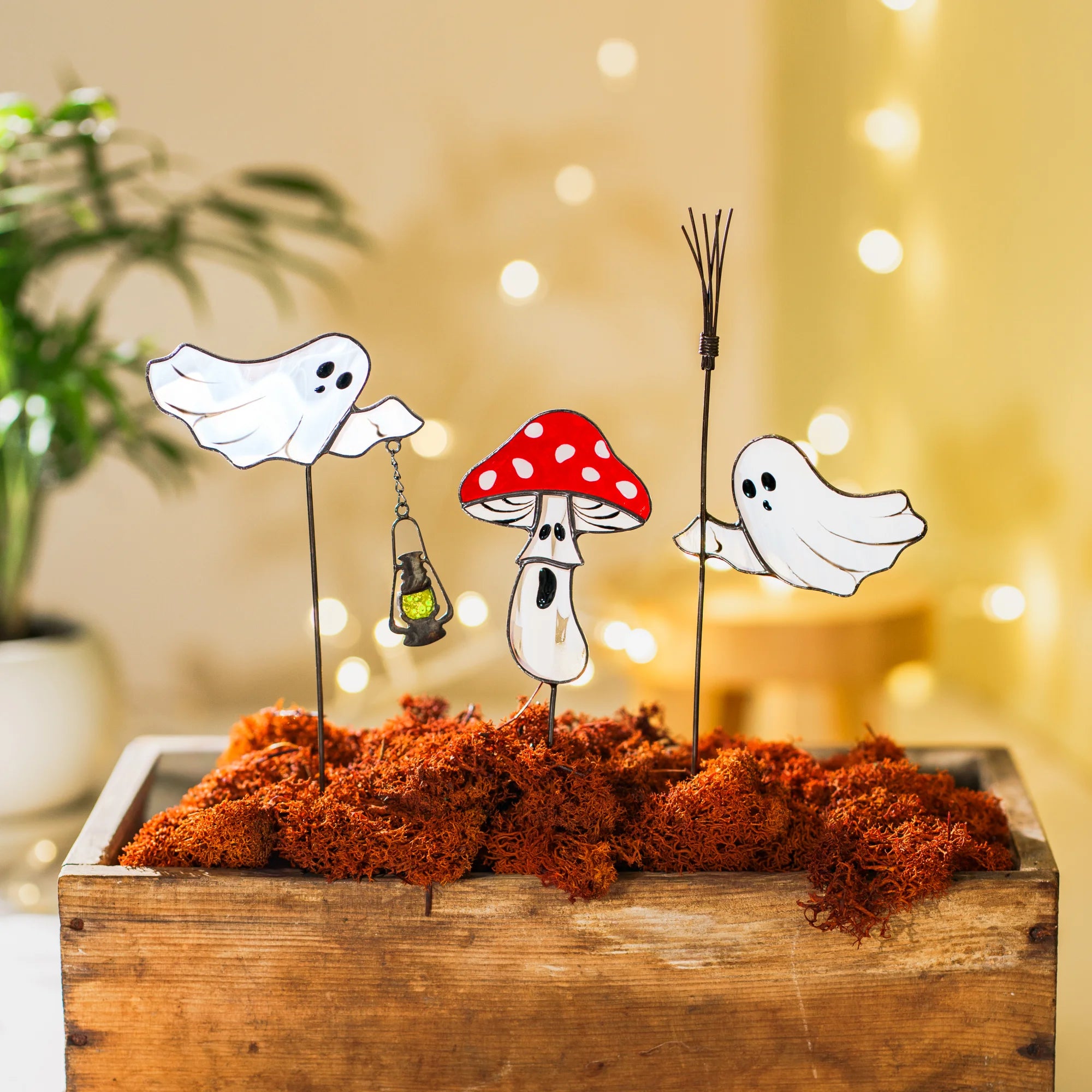 Set of three glass ghost and mushroom plant stakes in a wooden box of rust-orange moss, warm bokeh lights behind - moody stained glass halloween decor for a cozy autumn corner