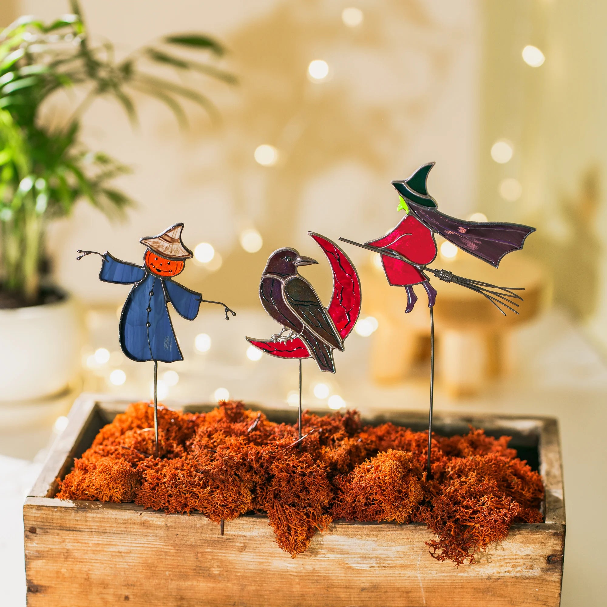 Scarecrow, raven and witch stained glass garden art staked in orange moss in a wooden box, warm bokeh lights behind - moody handmade Halloween decor for a cozy corner