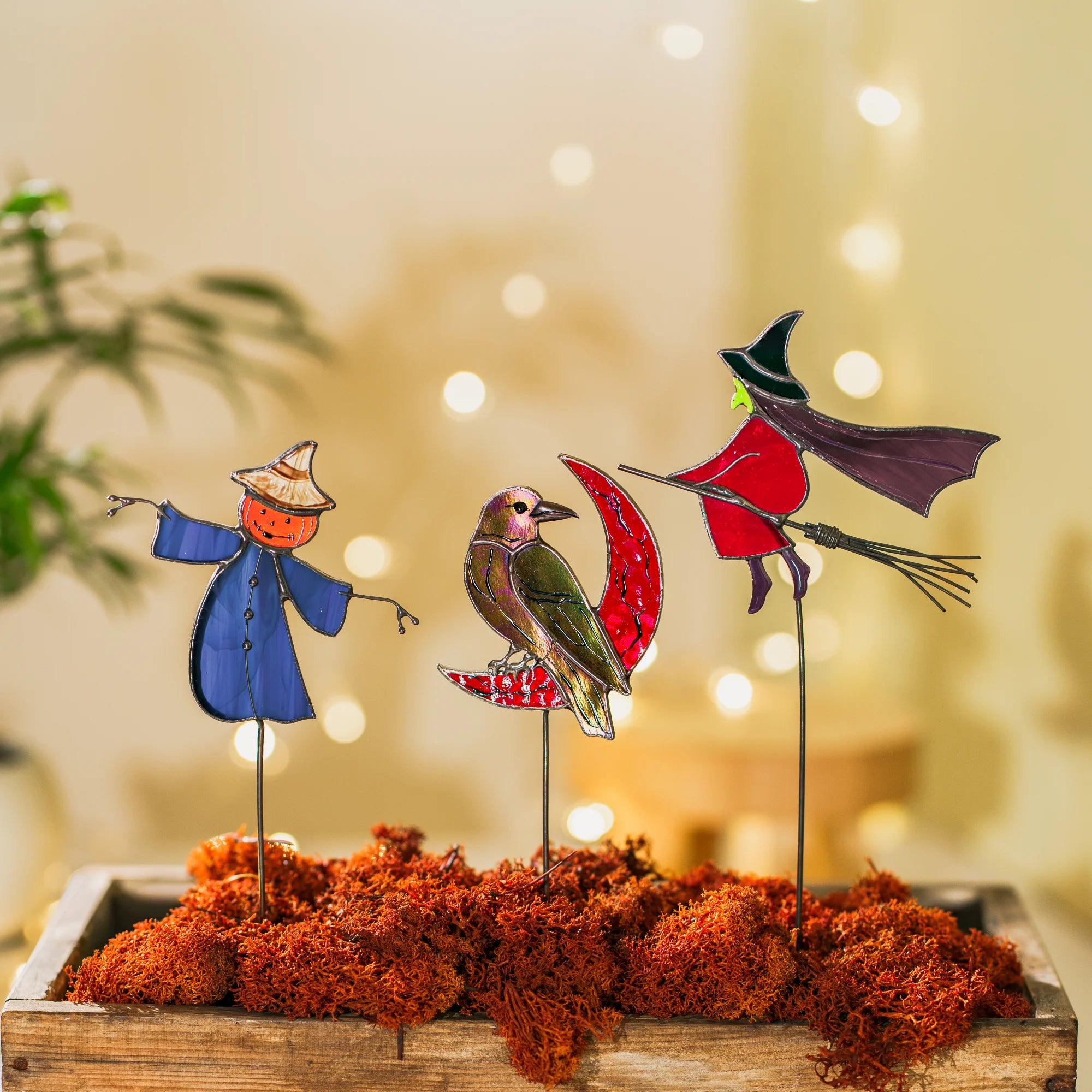 Close view of Halloween stained glass plant stakes in a mossy wooden box - iridescent raven on a red moon, green-faced witch on a broom and a grinning pumpkin scarecrow