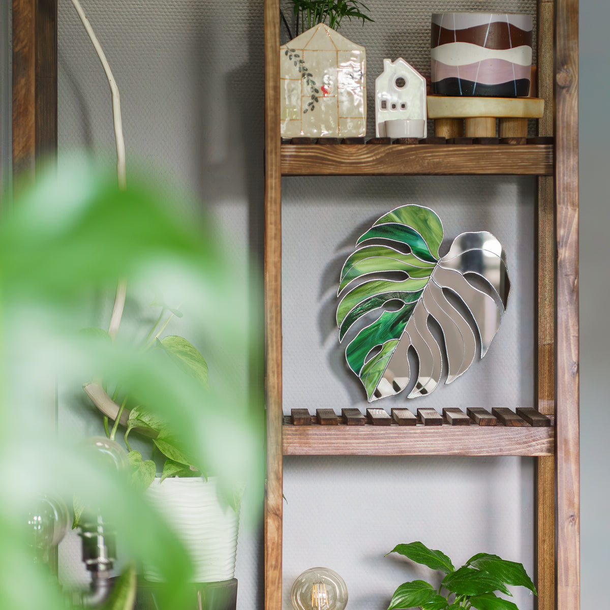 Monstera Leaf Stained Glass Mirror on a rustic wooden shelf seen through soft blurred greenery - handmade stained glass wall mirror and botanical decor, one of our favorite glass wall decor gifts