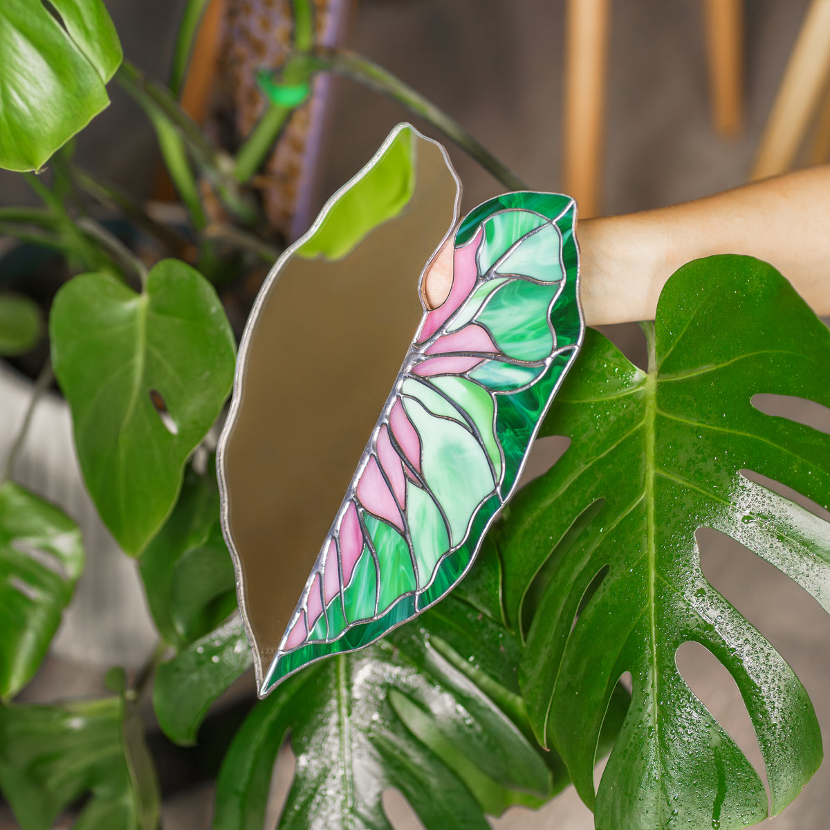 Close-up of the Caladium Leaf Stained Glass Mirror among dewy monstera leaves, showing swirled green and pink glass with soldered seams - handmade stained glass plants decor and wall art