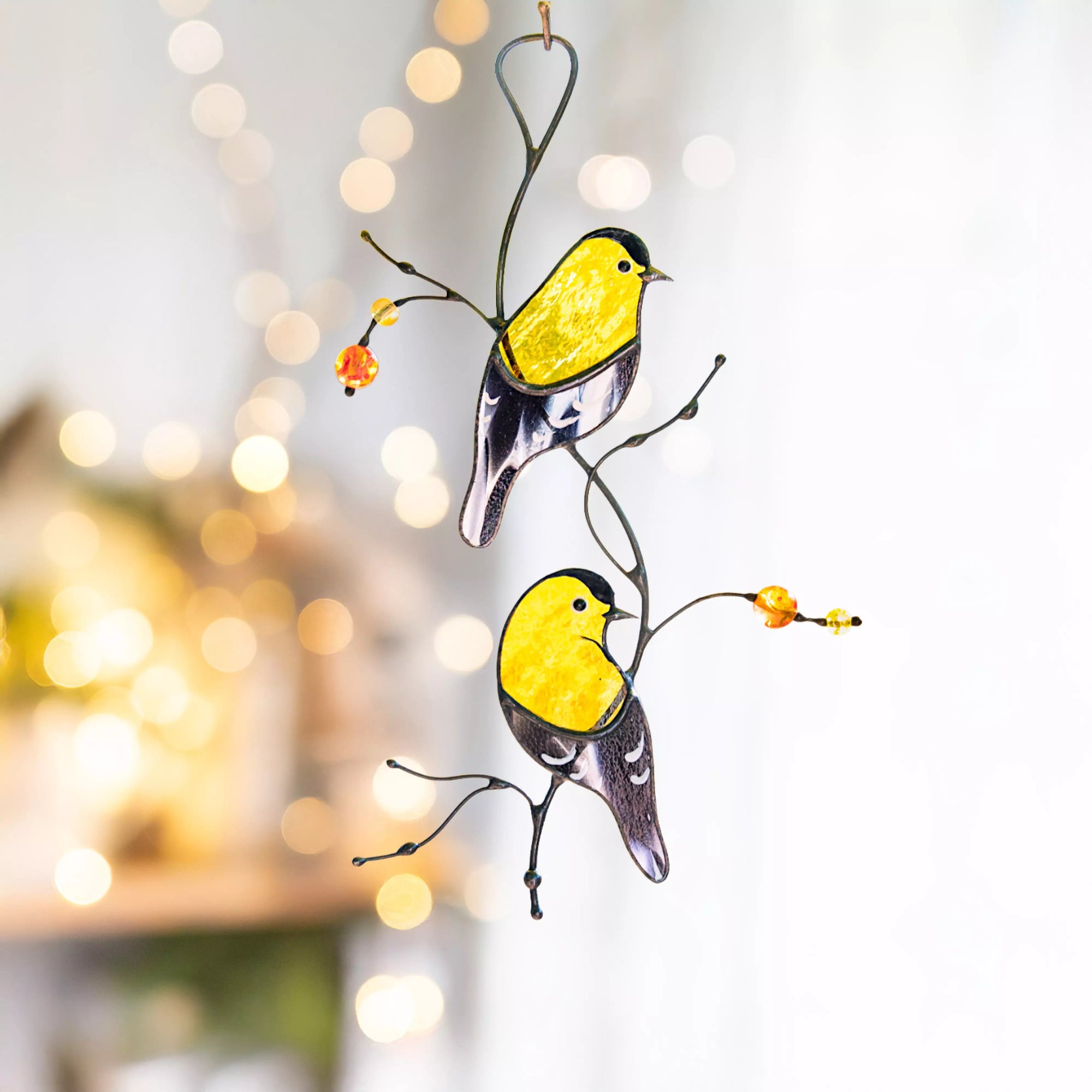 Lifestyle shot of the goldfinch suncatcher hanging indoors with soft bokeh lighting and indoor plants in the background.