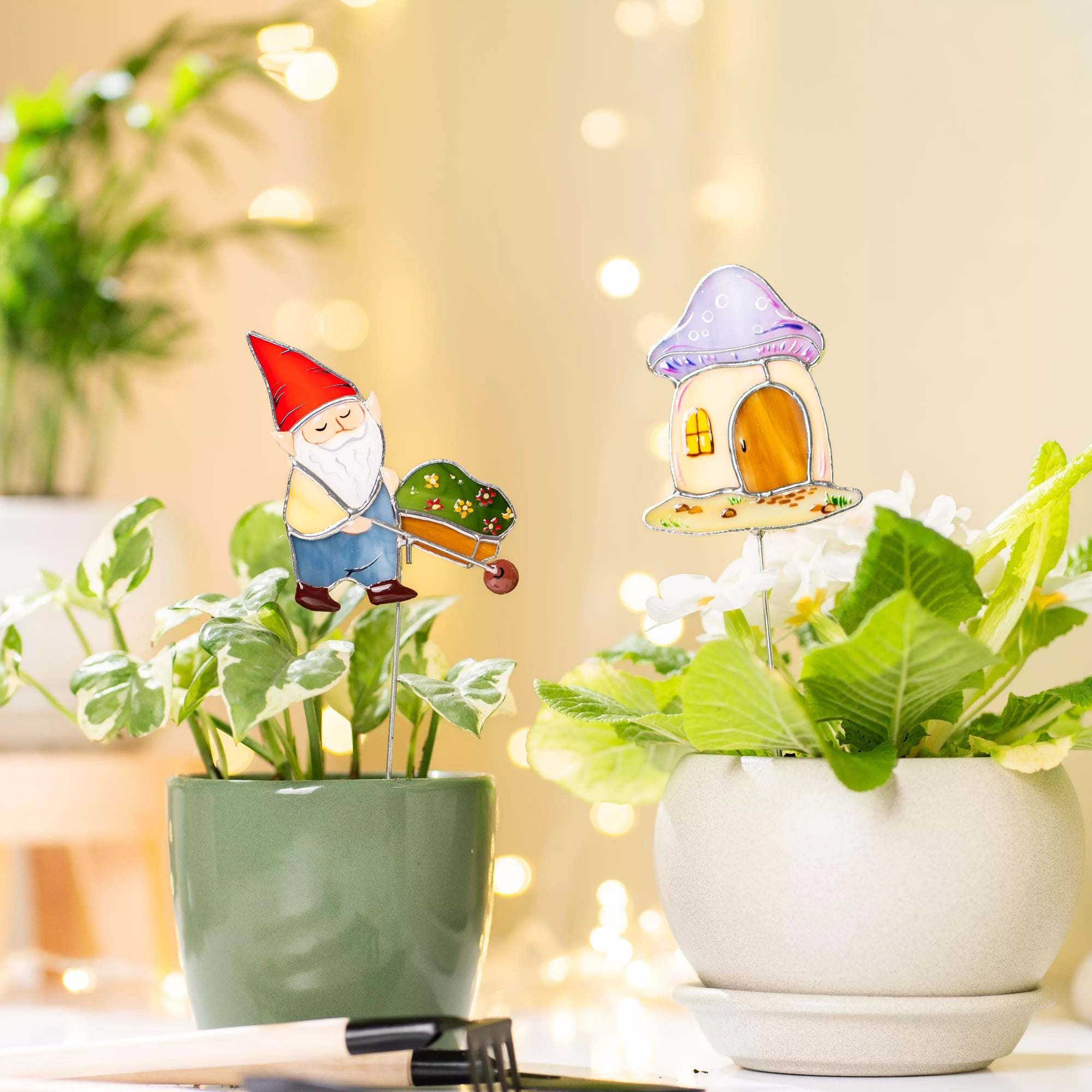 A farmhouse display featuring both garden stakes in their respective green and grey pots, set against a warm background of blurred fairy lights.