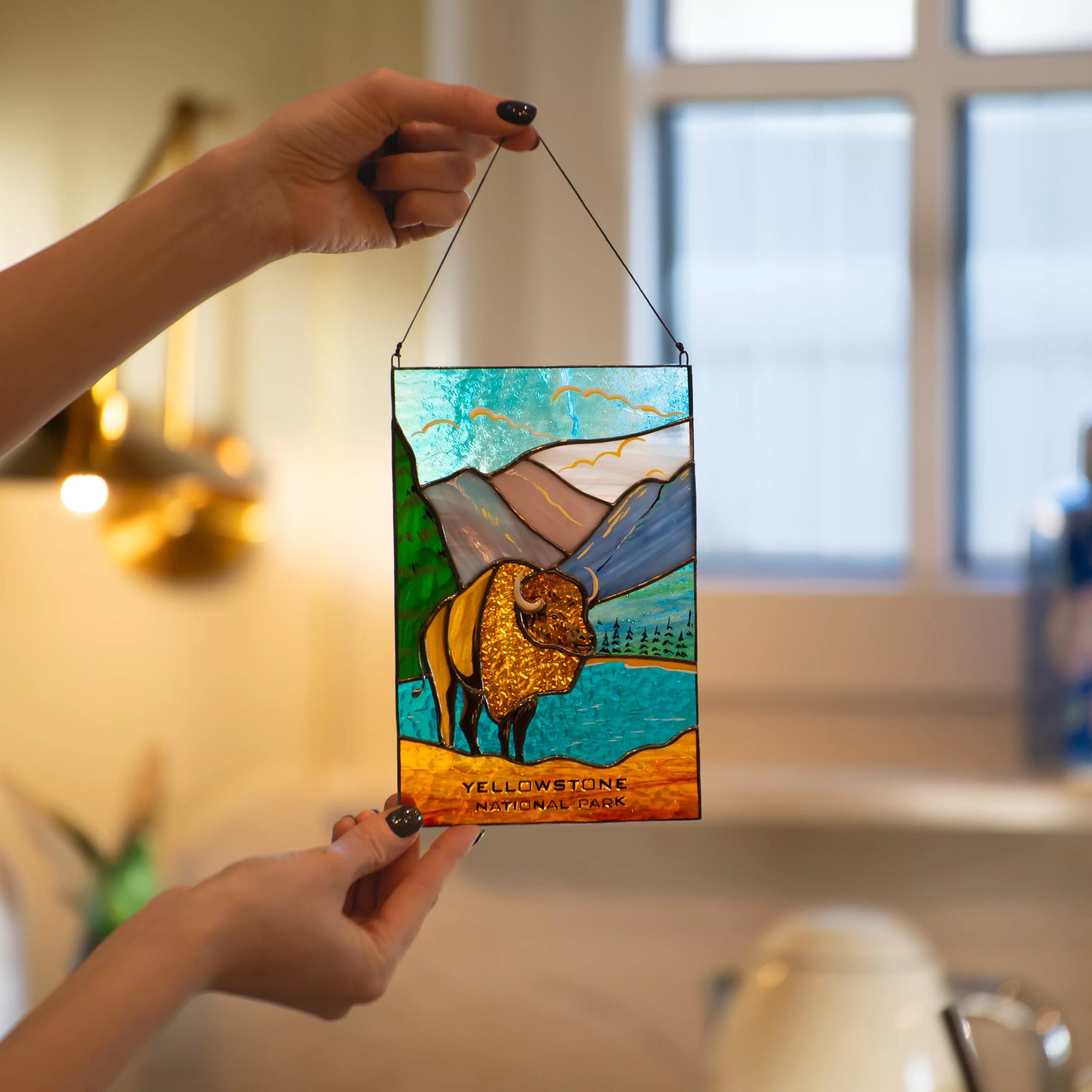 Hand holding a stained glass panel with a handshake design, featuring Yellowstone National Park, in a home setting.