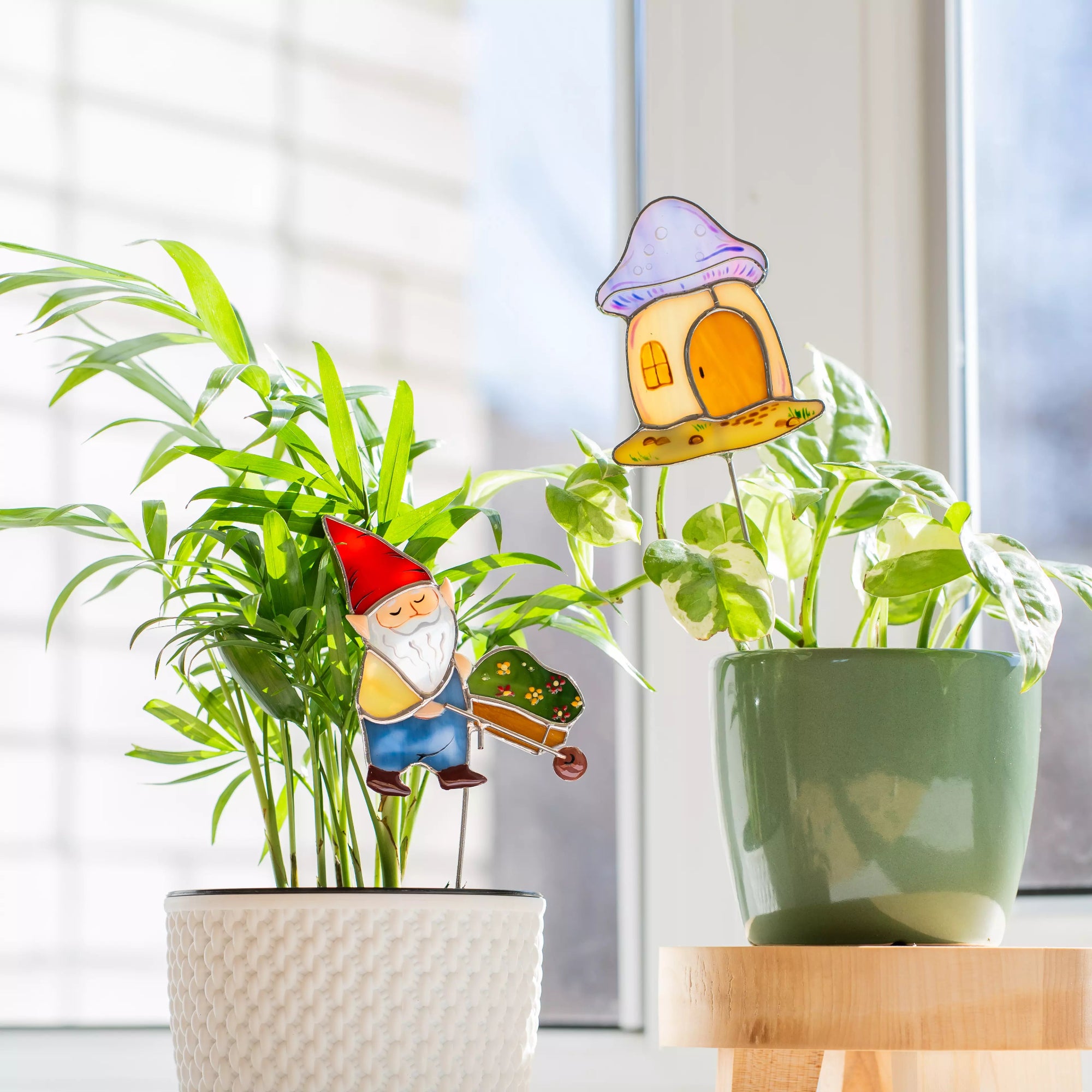 An indoor setting showing the gnome plant stake in a white pot on the left and the mushroom house garden stake in a green pot on a wooden stand to the right.