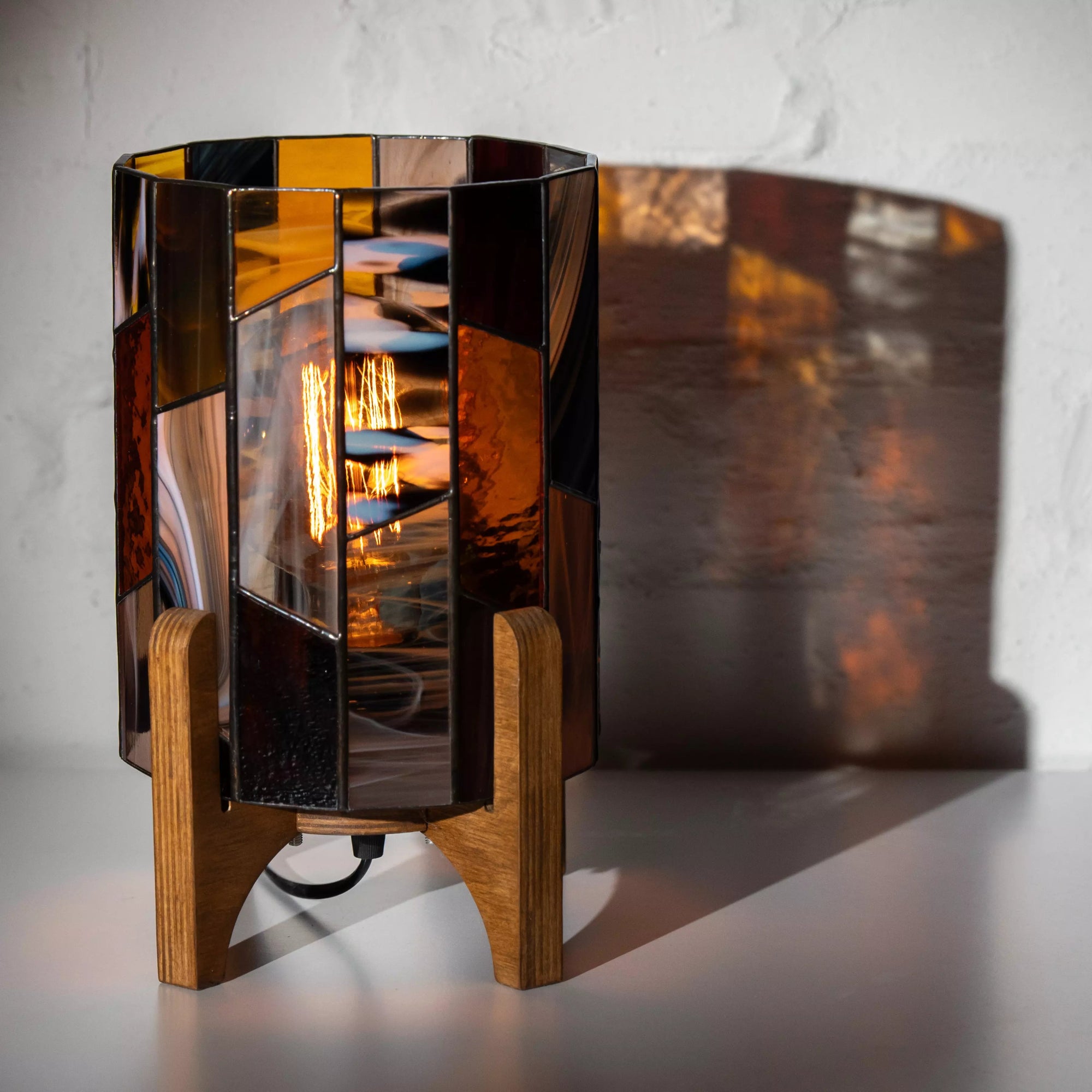 Handcrafted stained glass lamp casting warm reflections on the wall, amber and smoky glass design with wooden stand, perfect for modern interiors.