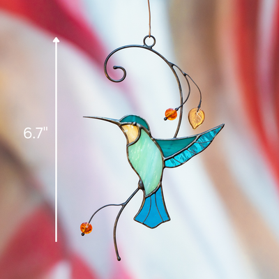 Hummingbird suncatcher with turquoise wings and hanging beads, displayed outdoors with soft red and beige background