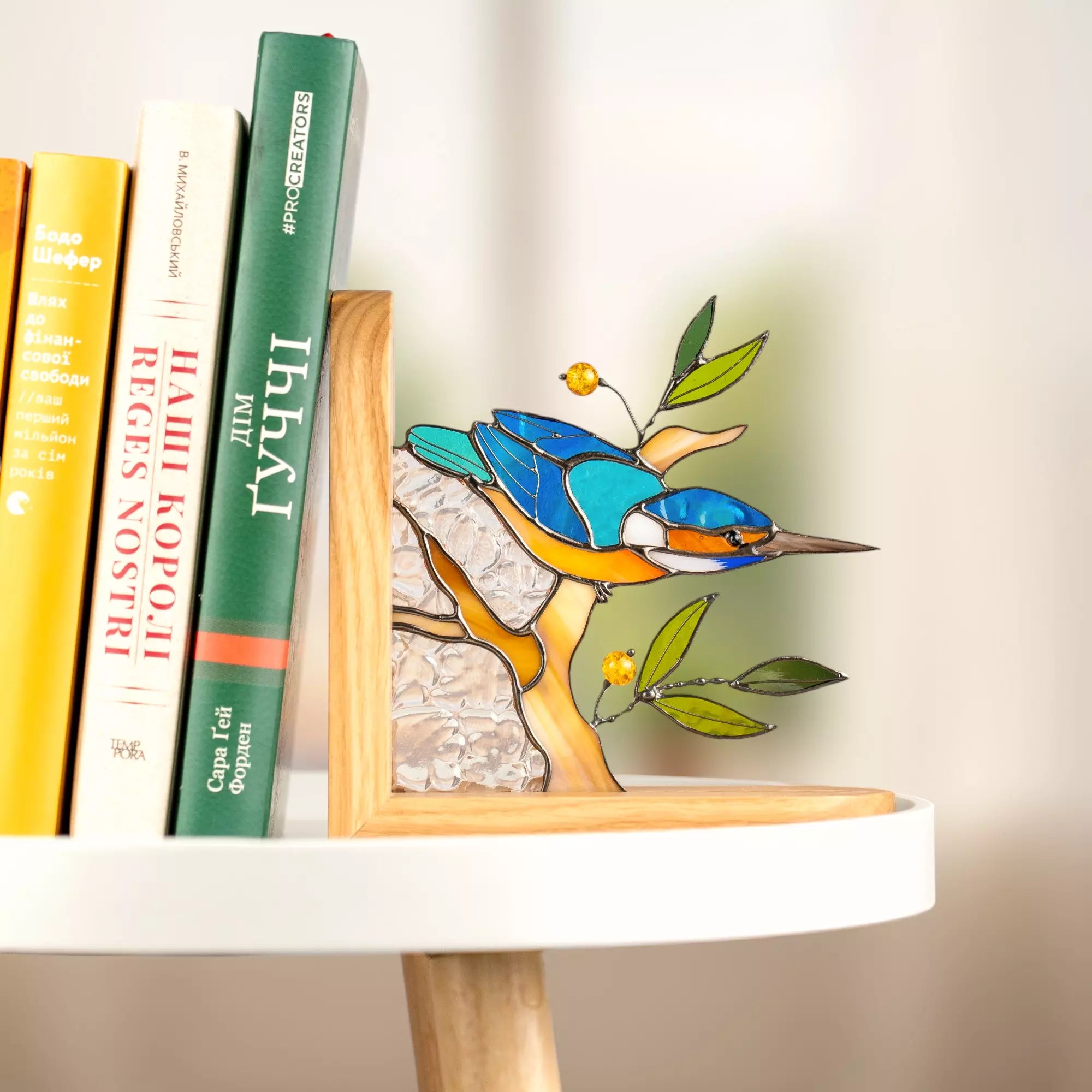 Modern stained glass kingfisher bird bookend displayed on a white round table. This piece combines natural oak wood with artisan glasswork, perfect for contemporary interior design. 