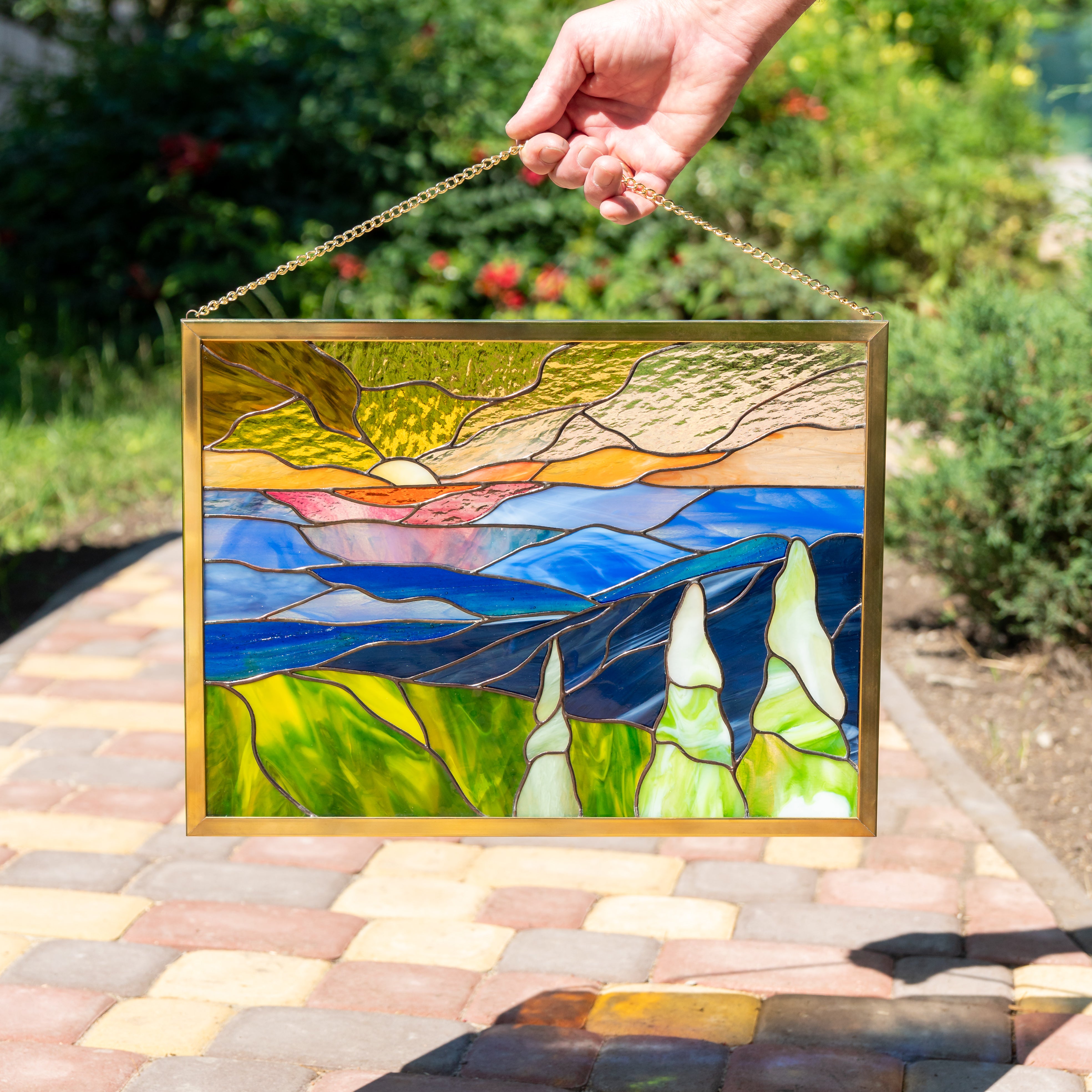 Hand holding stained glass panel outdoors with trees and path in the background