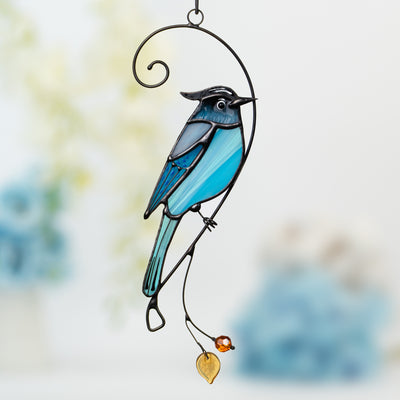 Stained glass Steller’s jay in vivid blue tones with decorative branch, amber bead, and yellow leaf hanging below