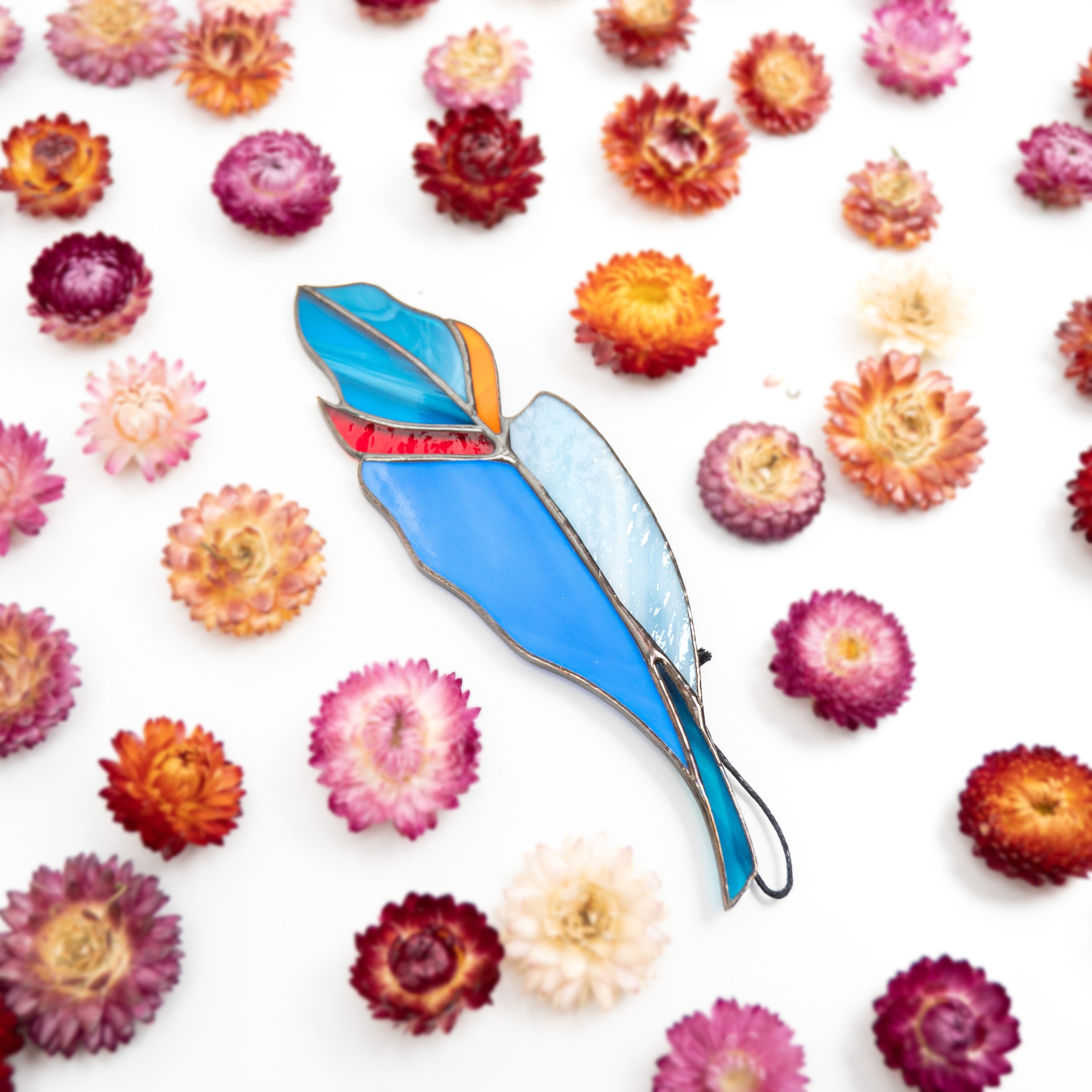 Multicolored stained glass feather laying among scattered strawflowers in purple, yellow, and pink tones.