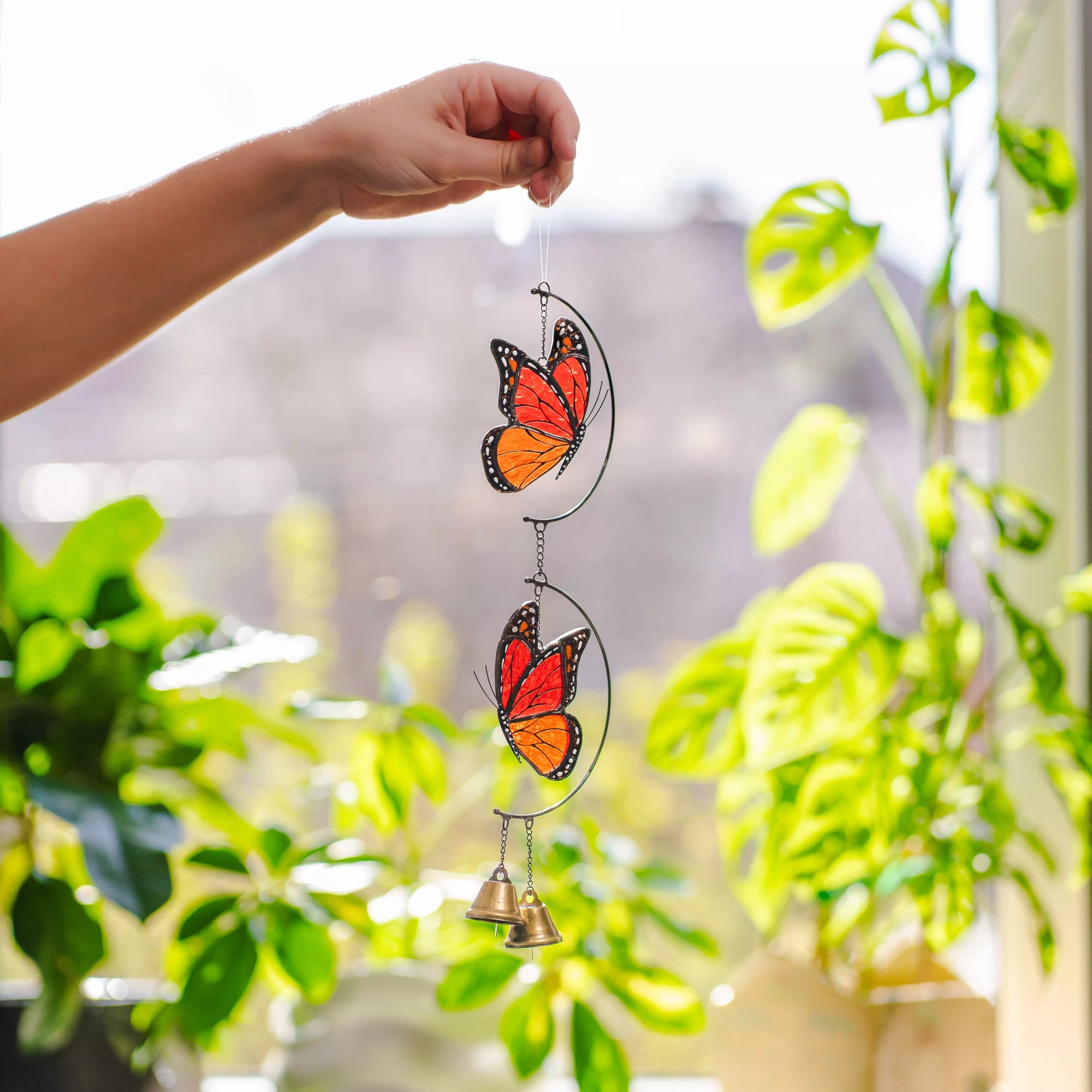 Butterfly stained glass wind chime with a bell held by a hand in front of green plants.