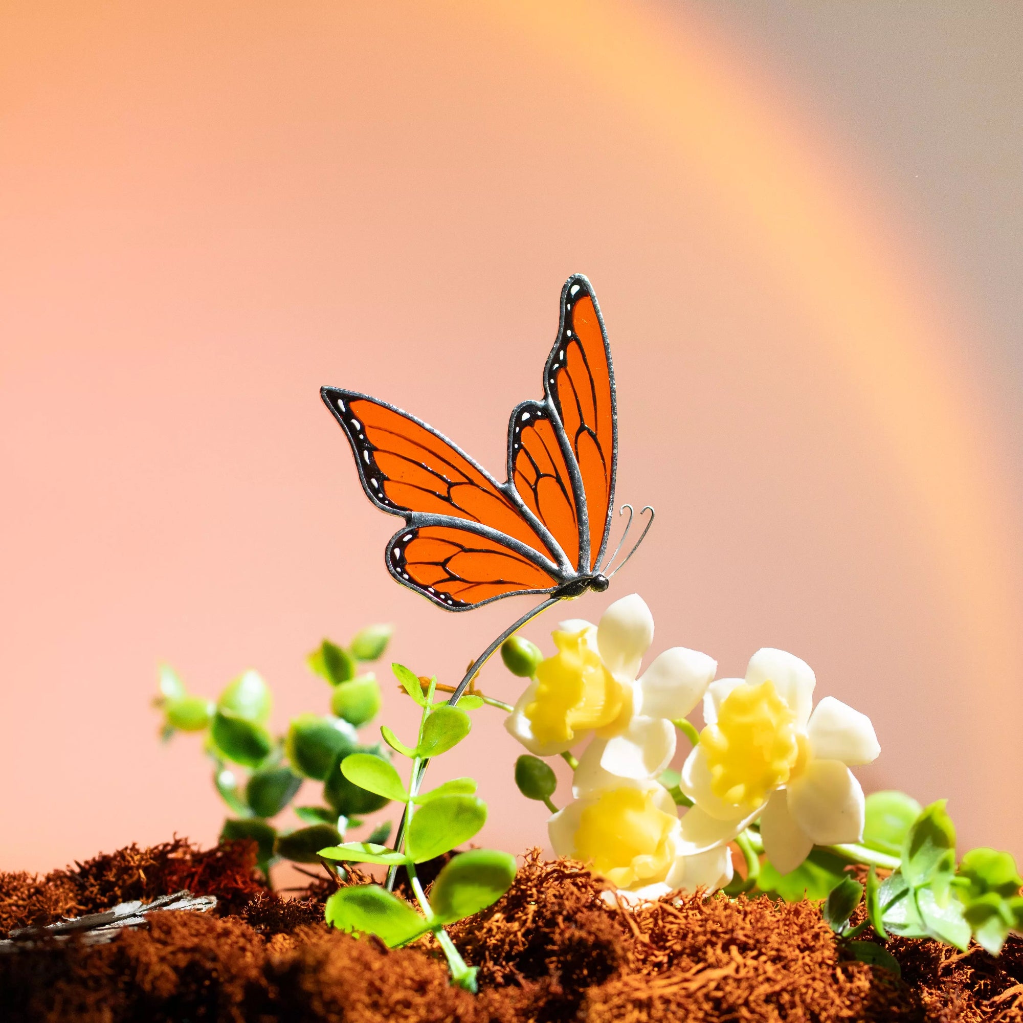 Orange stained glass butterfly stake placed in soil with small yellow flowers and green plants.