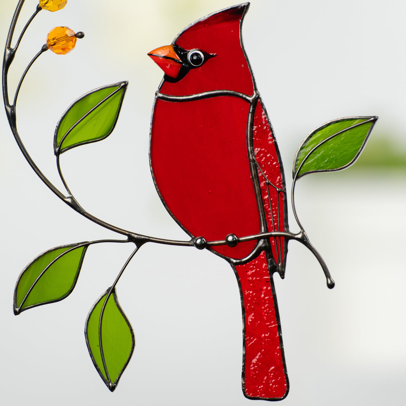 Close-up of stained glass red cardinal sitting on a curved branch with green glass leaves.