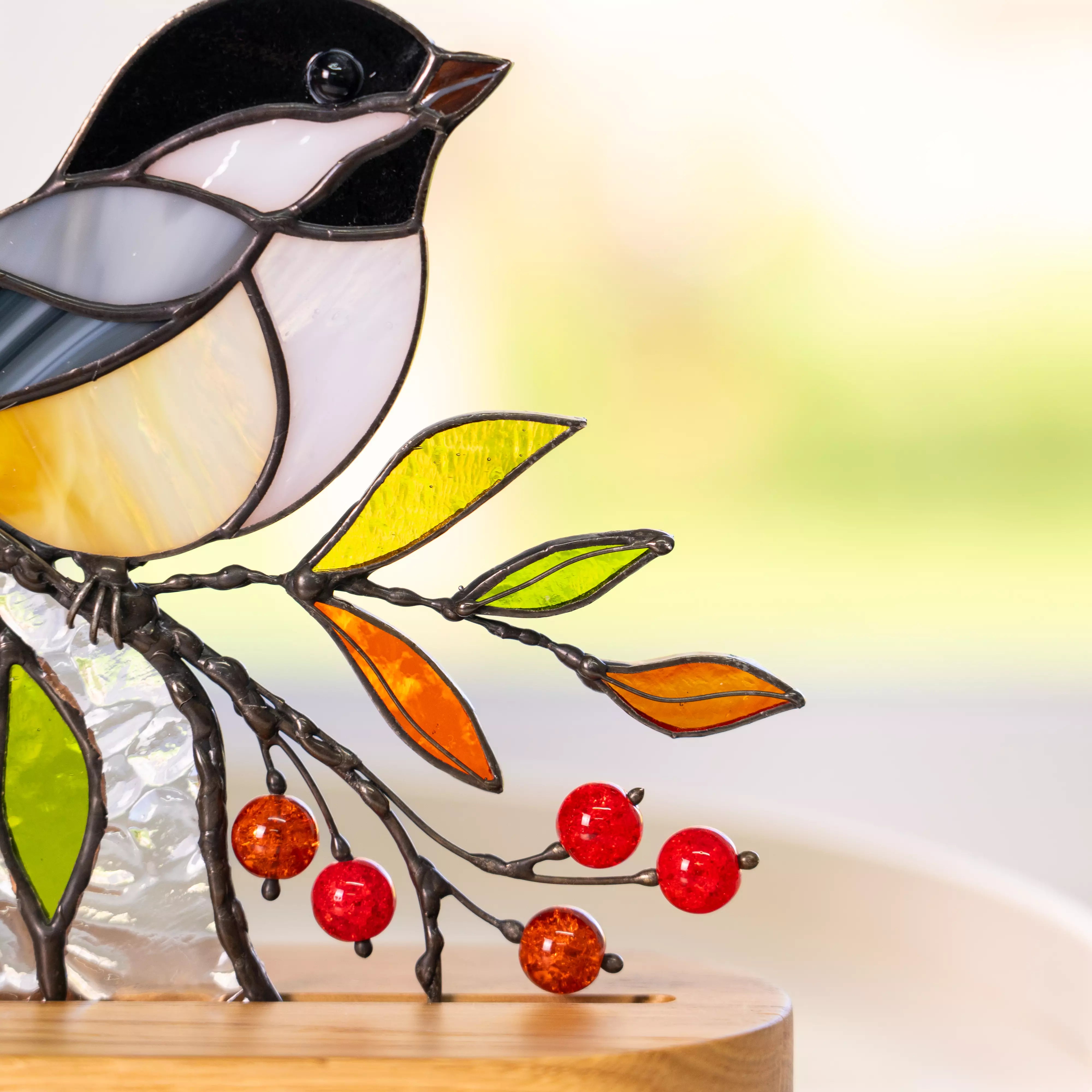 Close-up of stained glass chickadee with black cap, white cheeks, and yellow belly, surrounded by red glass beads and autumn leaves.