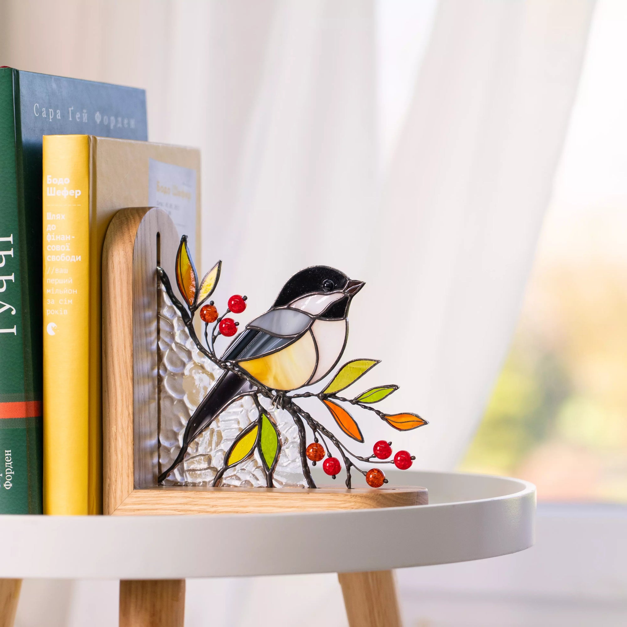 Stained glass chickadee bookend displayed on a white table near books, featuring red berries, colorful leaves, and a natural wooden frame.