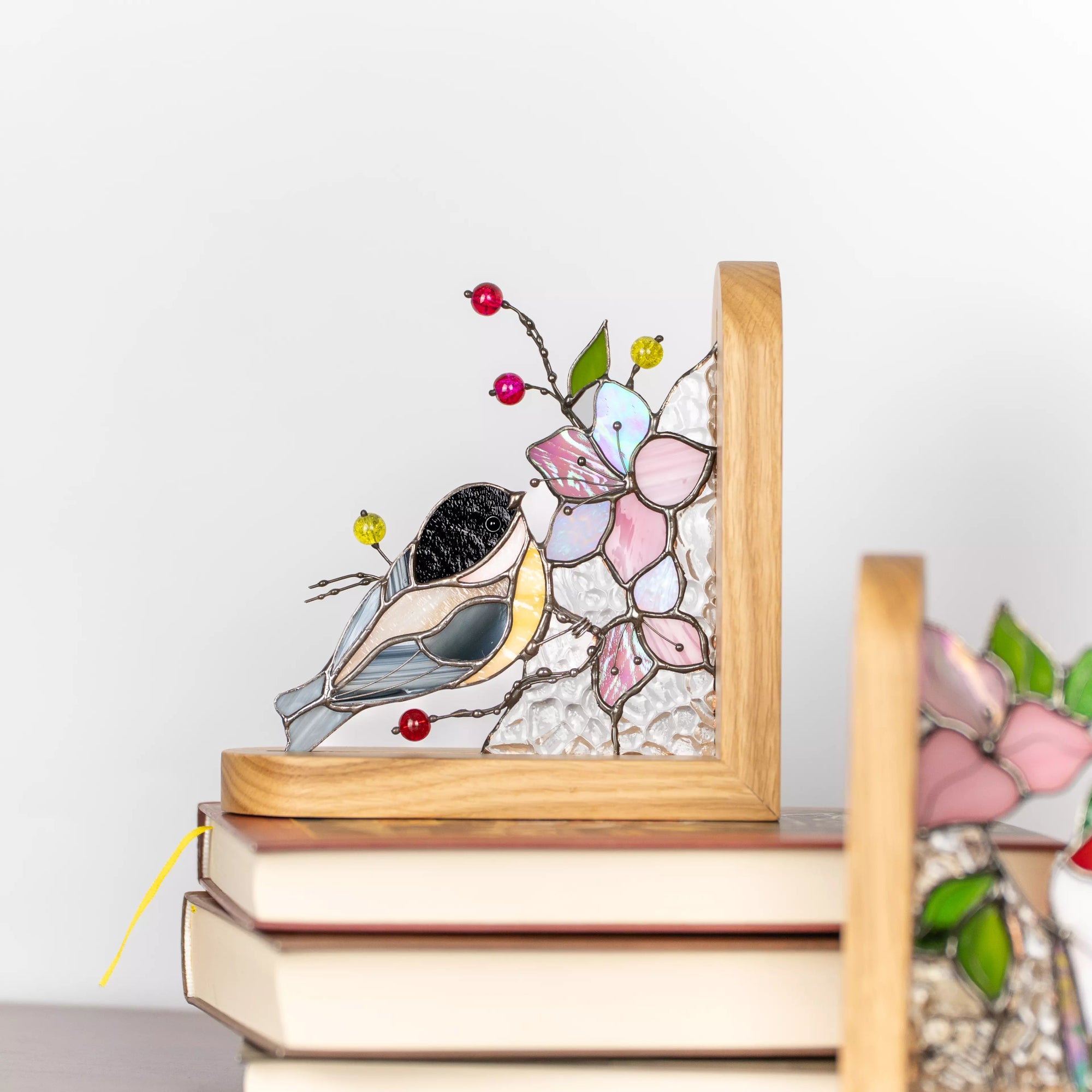 Stained glass bird bookend displayed on stacked books, featuring a chickadee, pink blossoms, textured clear glass, and natural wooden frame.