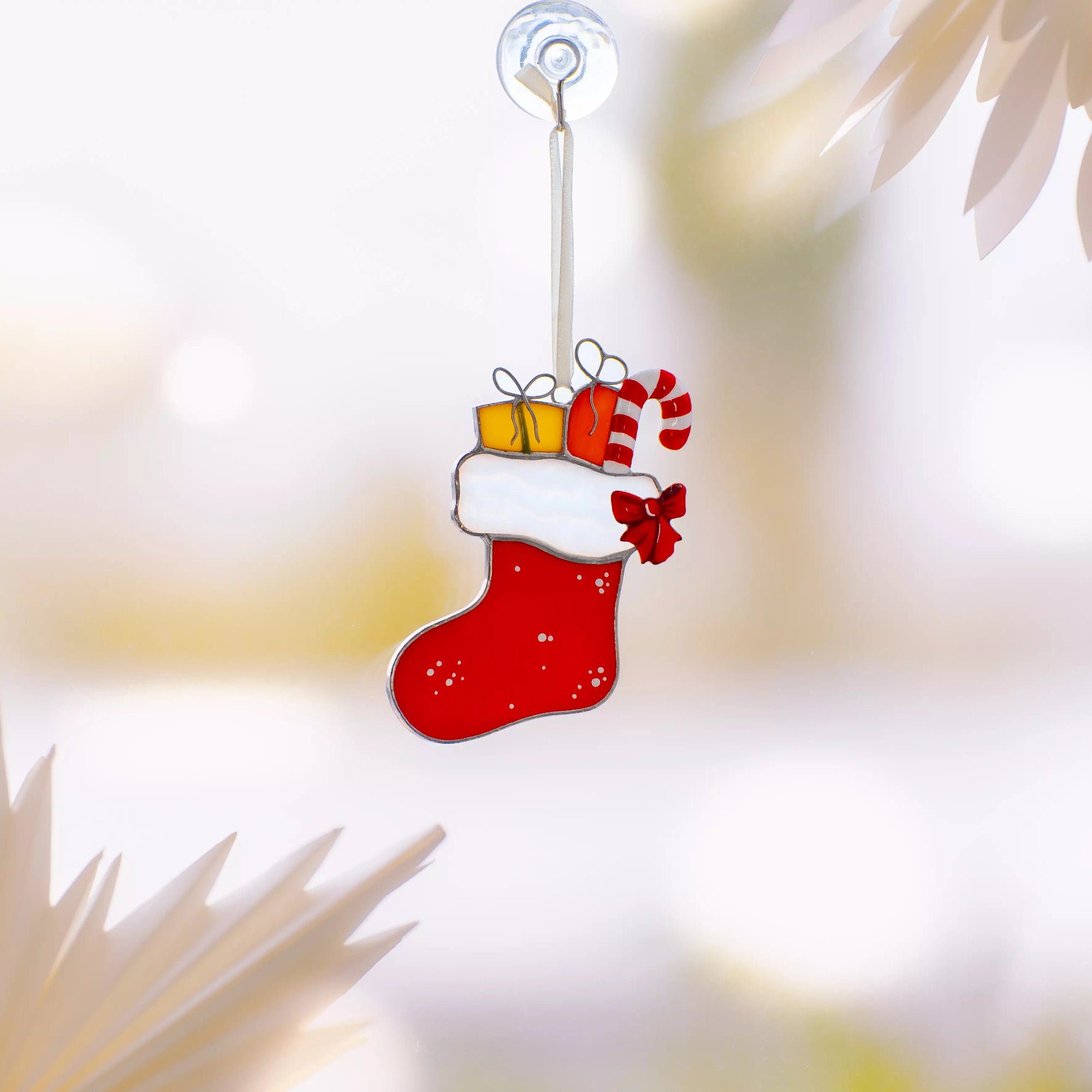 Red stained glass Christmas stocking with white top, gifts, and candy cane hanging on a suction cup in bright window light.