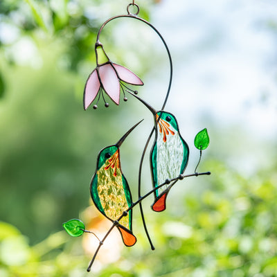 Stained glass green hummingbirds suncatcher hanging outdoors in sunlight near green foliage.