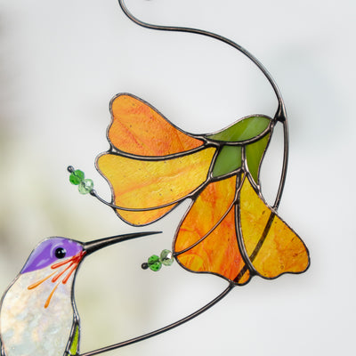 Detailed view of stained glass orange flower with green accents and a hummingbird beak nearby